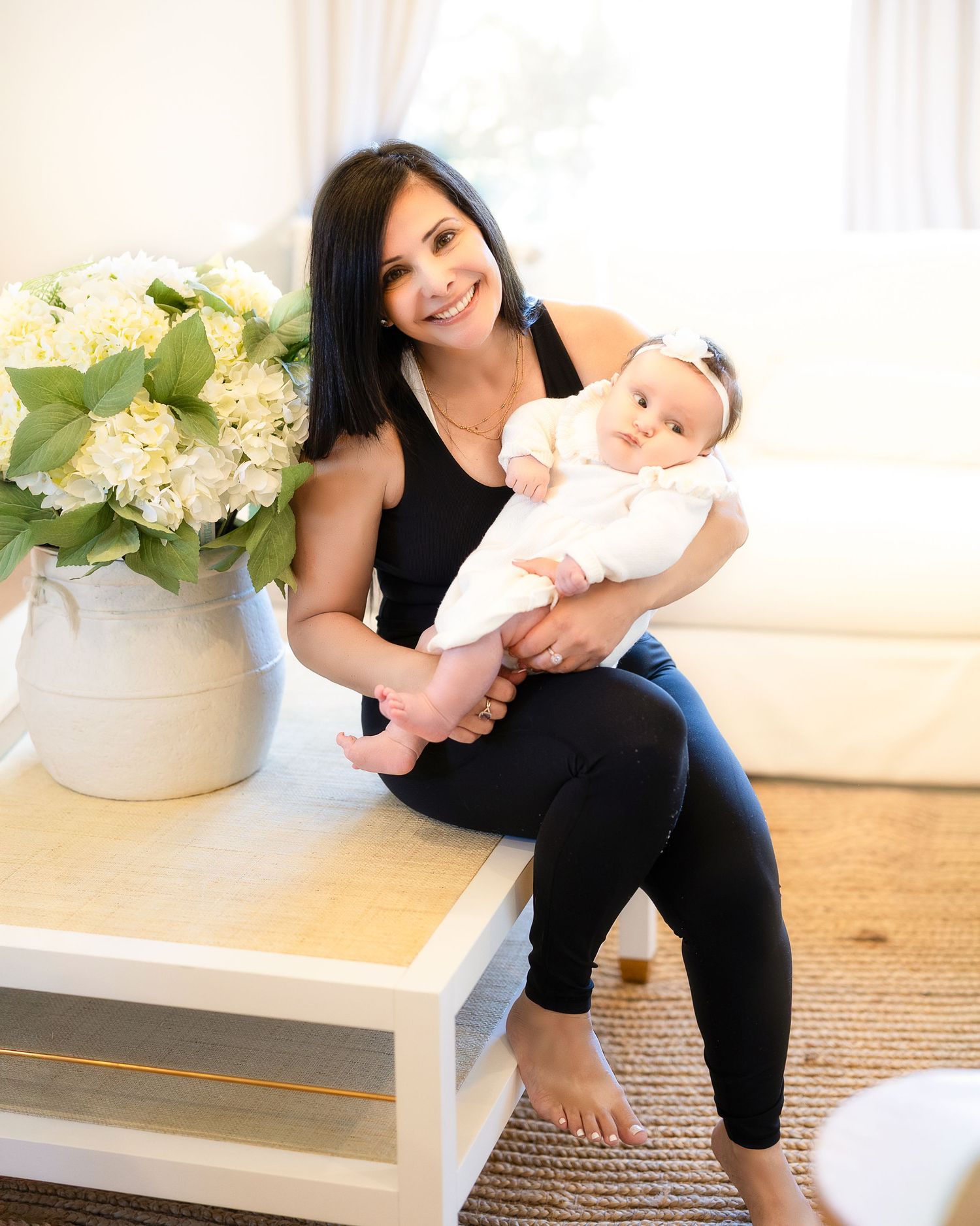 A woman in black workout clothes sits on a white bench with an infant in a pink outfit.