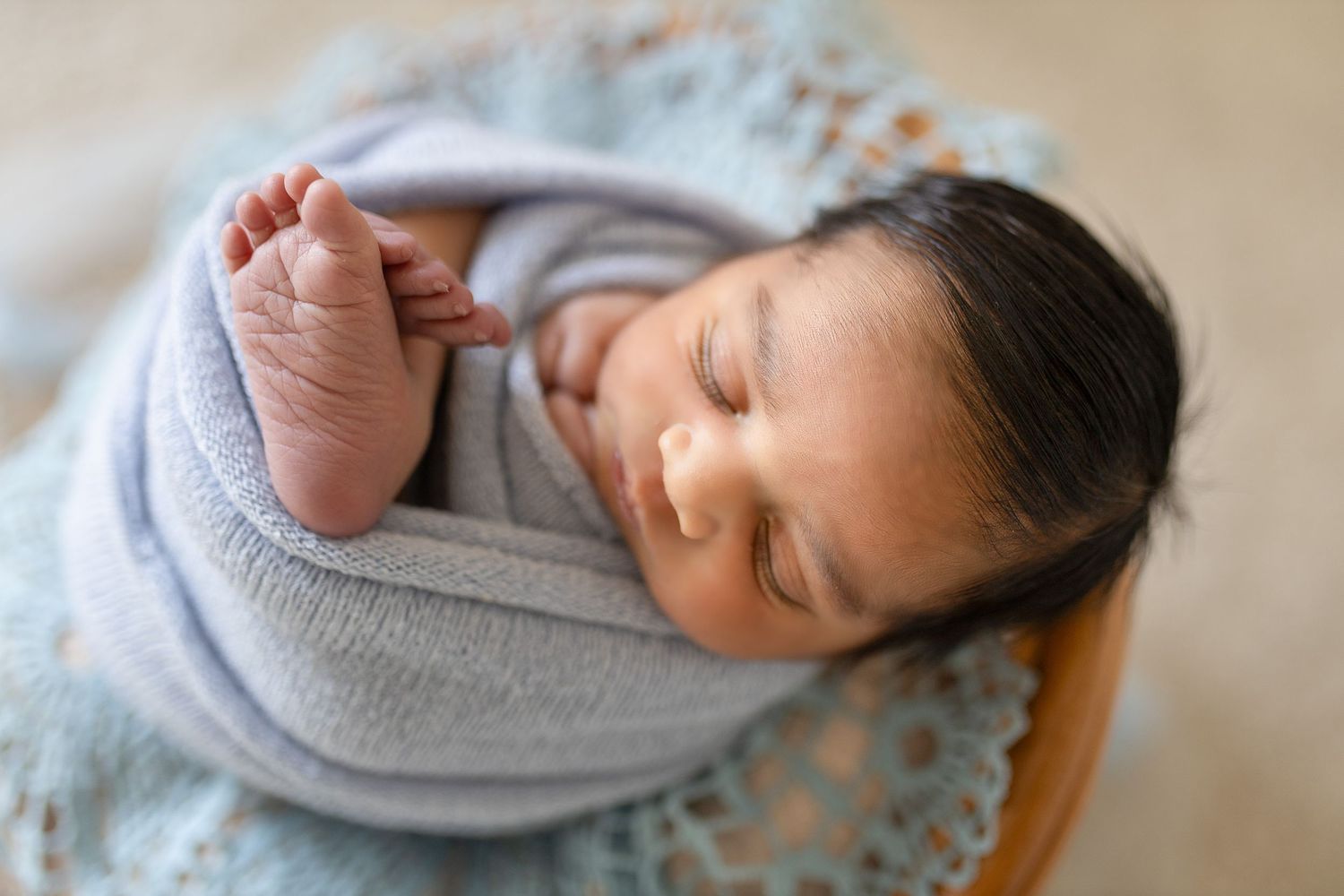 Sleeping baby swaddled in gray blanket showing tiny foot while resting on blue lace surface.