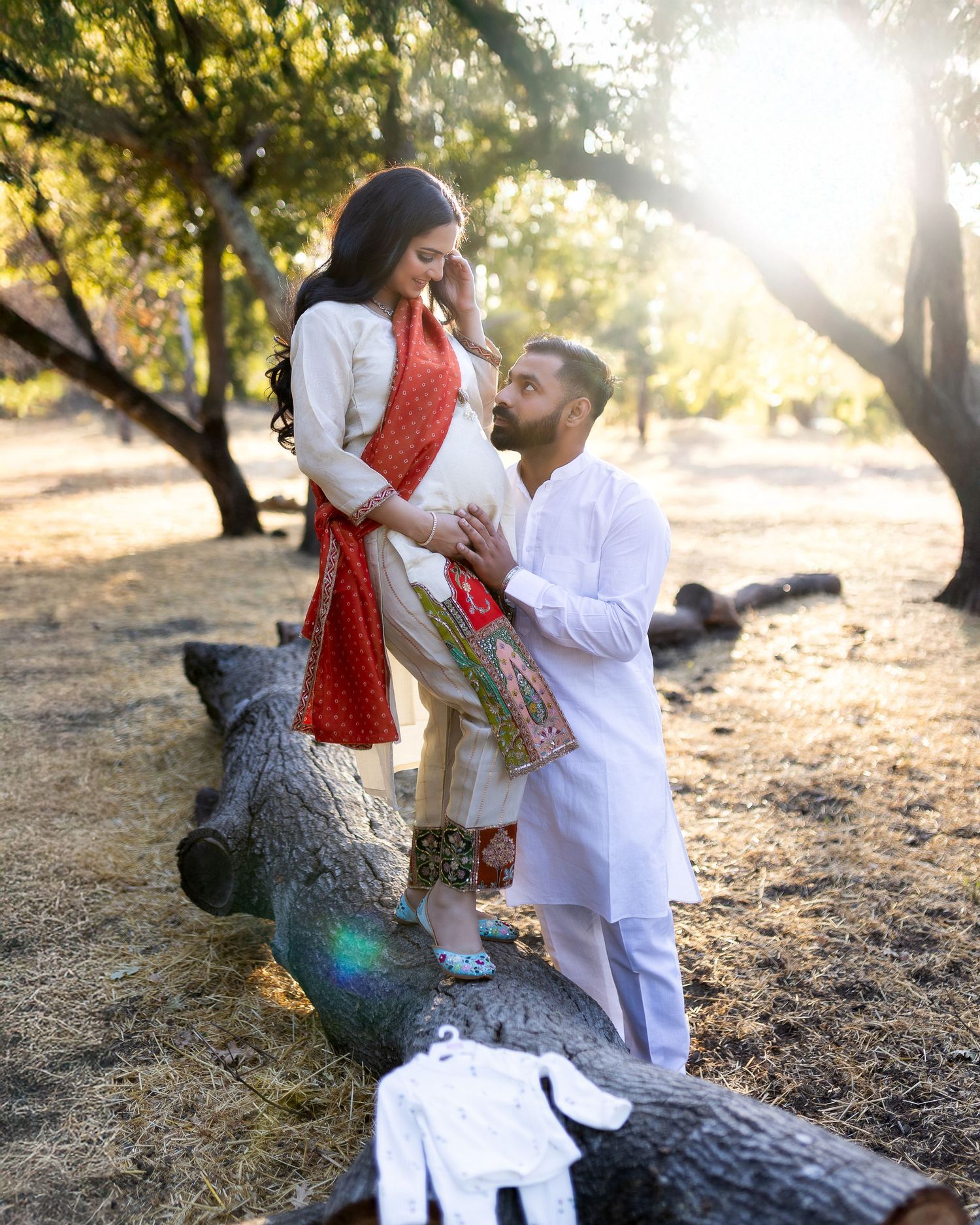A couple shares a romantic moment while sitting on a fallen log in a sunlit forest setting during golden hour.