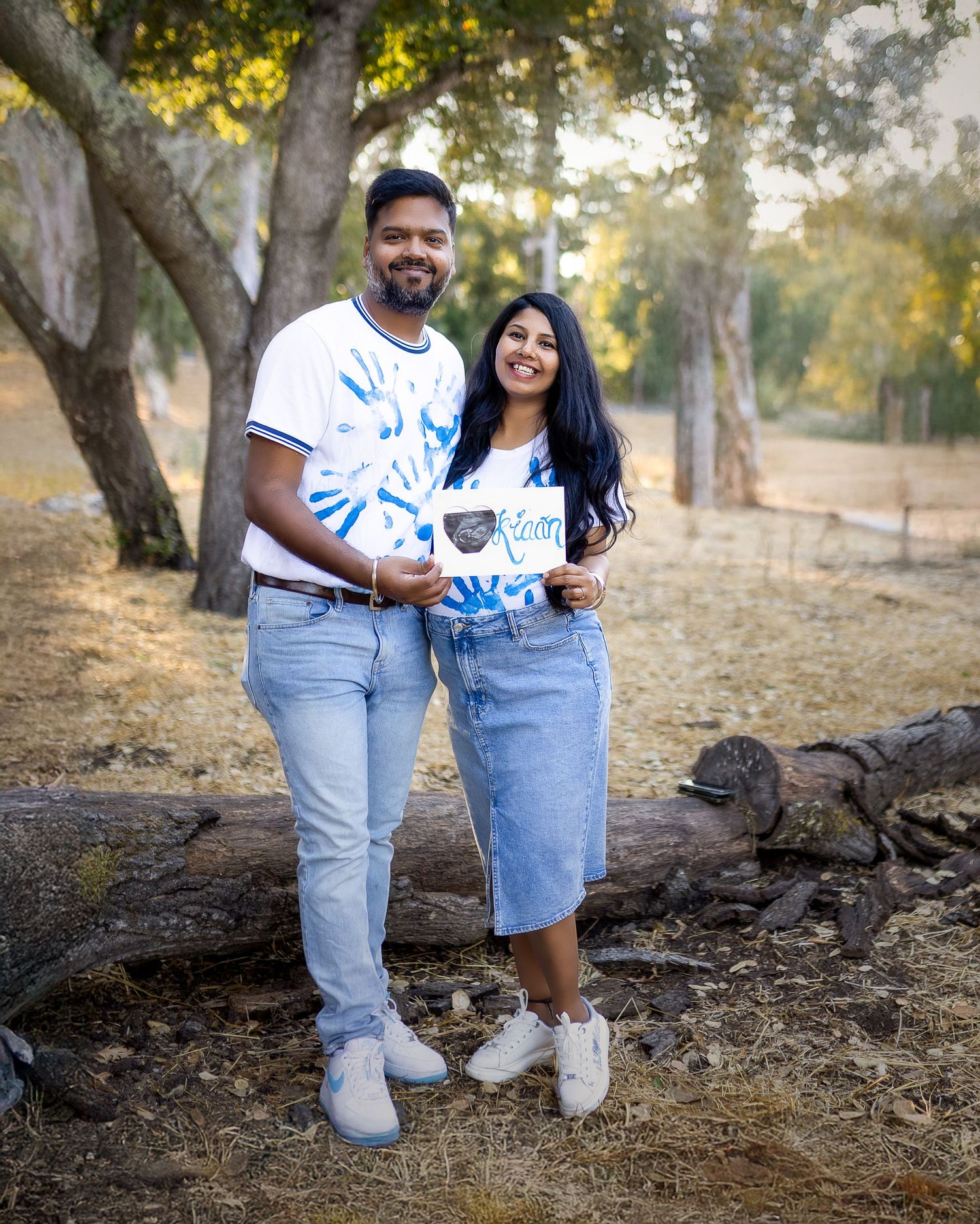 A couple in matching tie-dye shirts and jeans poses together outdoors among trees in a natural setting.