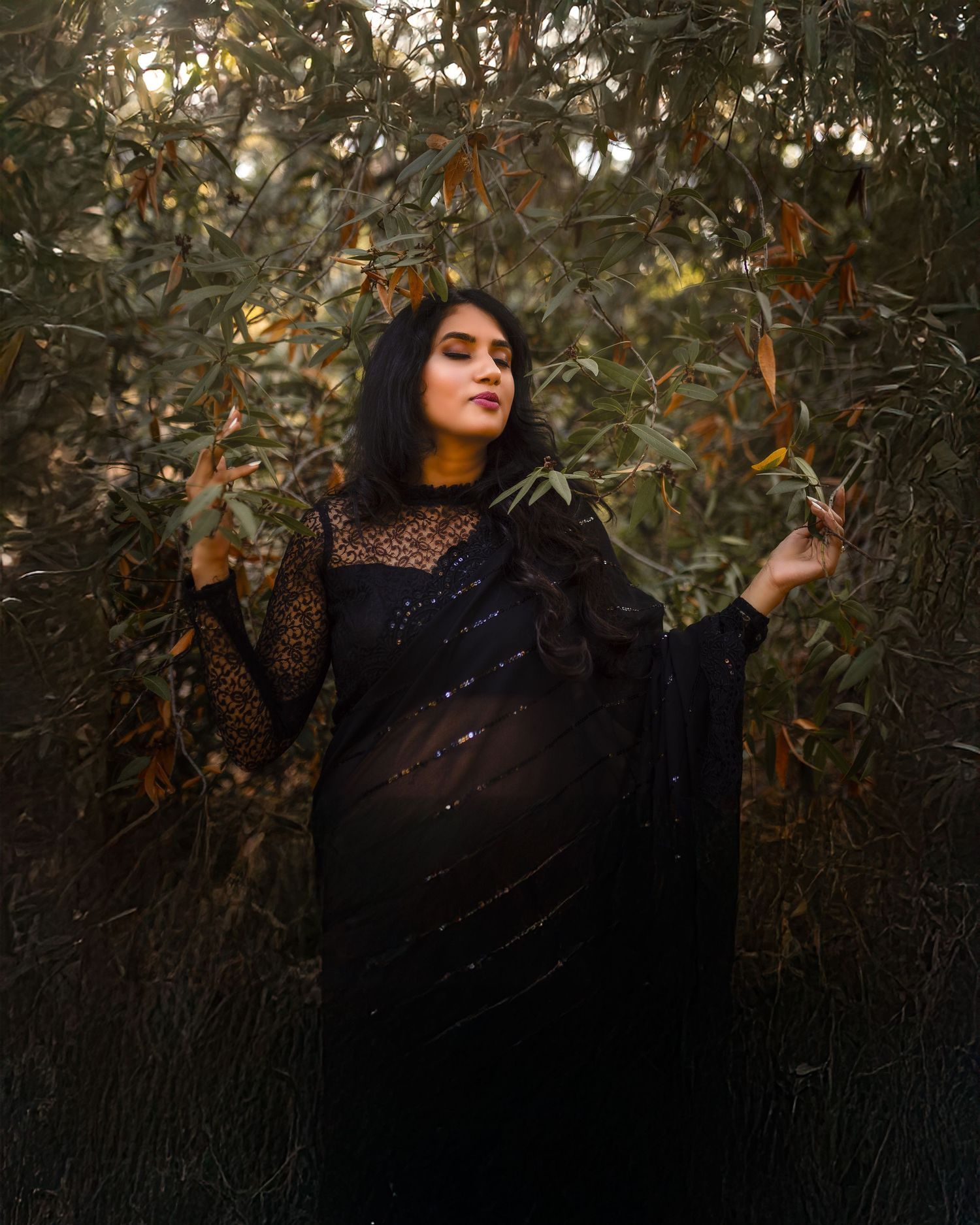 A person in a black outfit poses artistically among autumn foliage in a moody outdoor photoshoot.