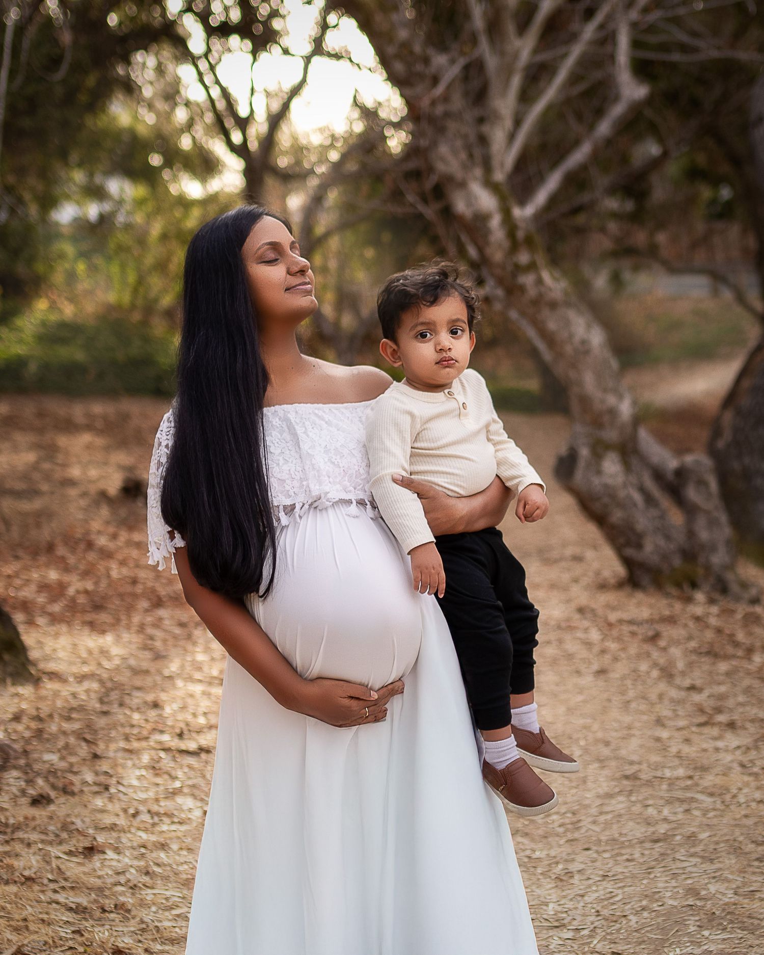 Mother and child share tender moments in white dress against natural outdoor backdrop during golden hour photography session.