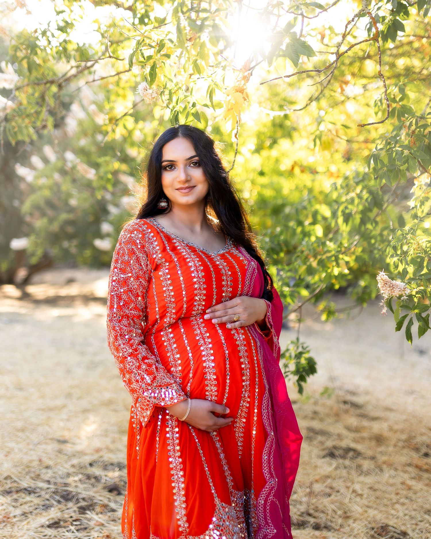 Maternity photo session in golden sunlight showing woman in vibrant red traditional dress with pink dupatta.