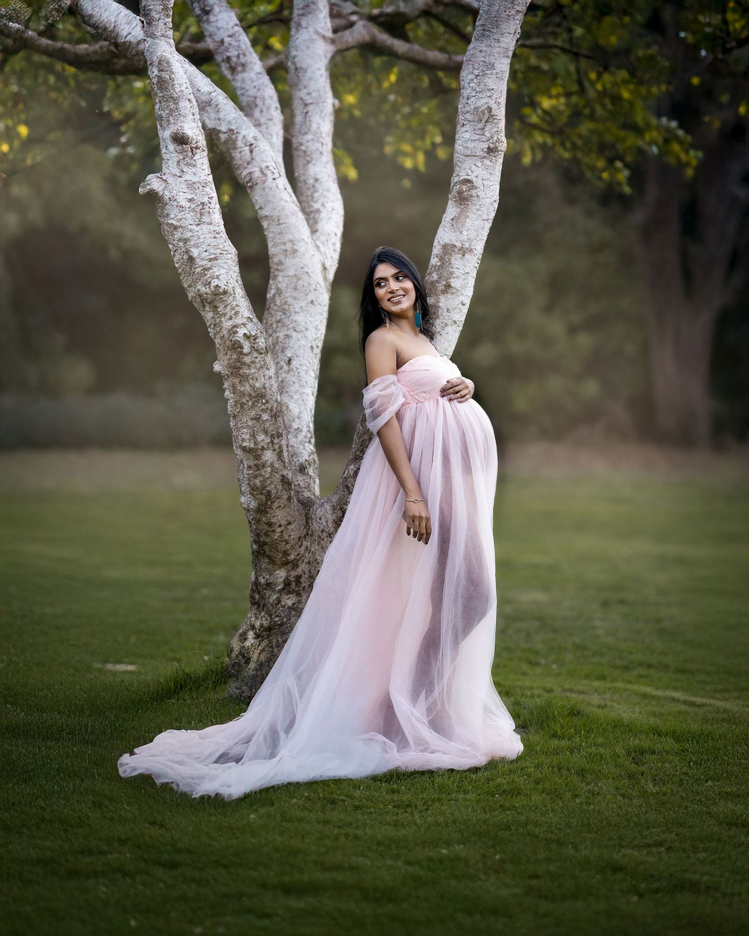An ethereal maternity photo featuring a pink tulle gown against a white birch tree in a dreamy outdoor setting.
