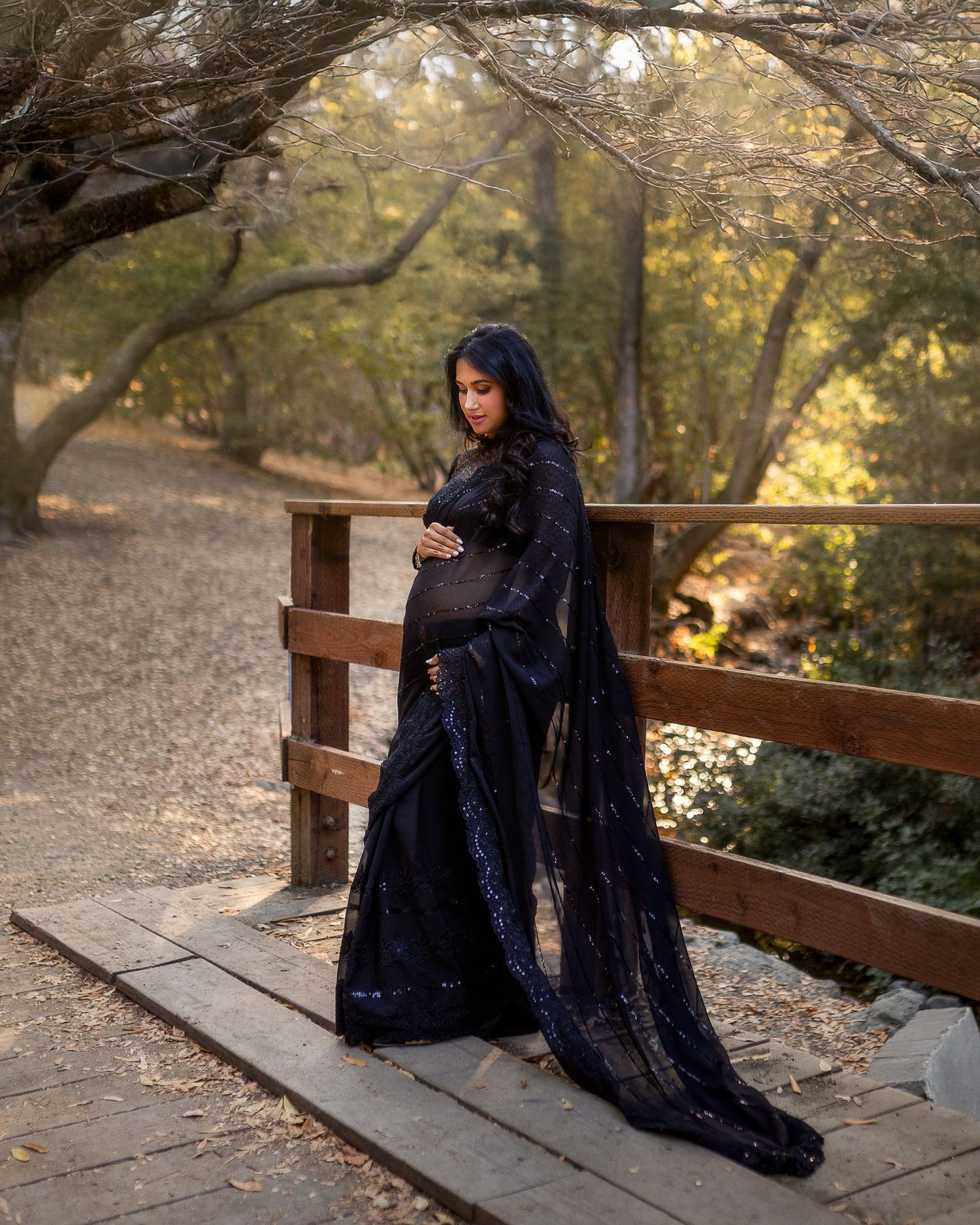 A figure in a flowing black dress sits contemplatively on a wooden bridge in a serene autumn forest setting.