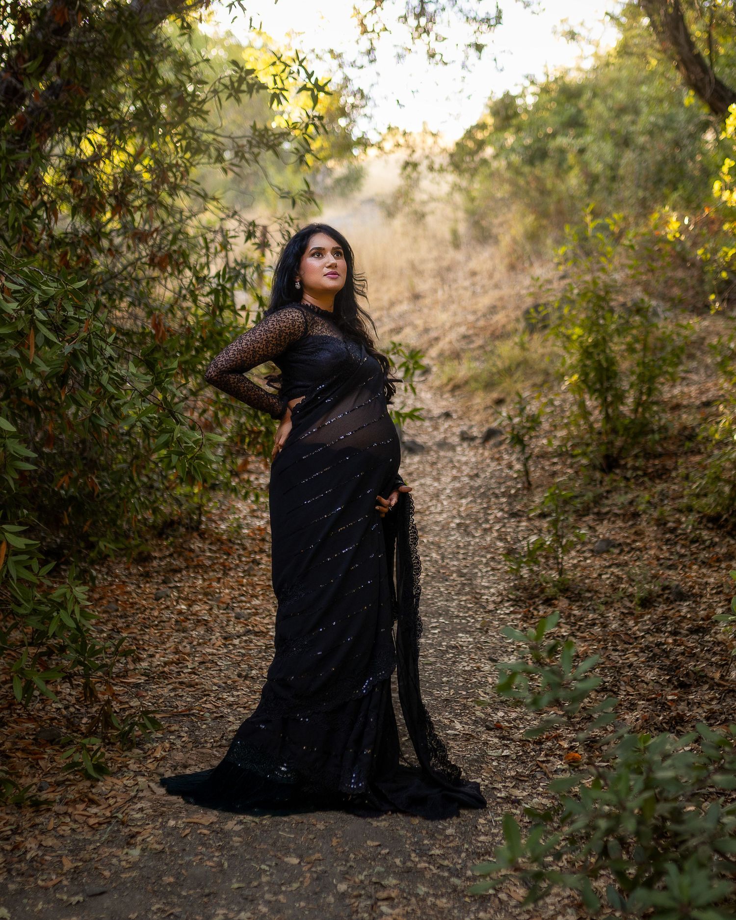 A figure in a flowing black dress poses gracefully on a wooded path during golden hour.