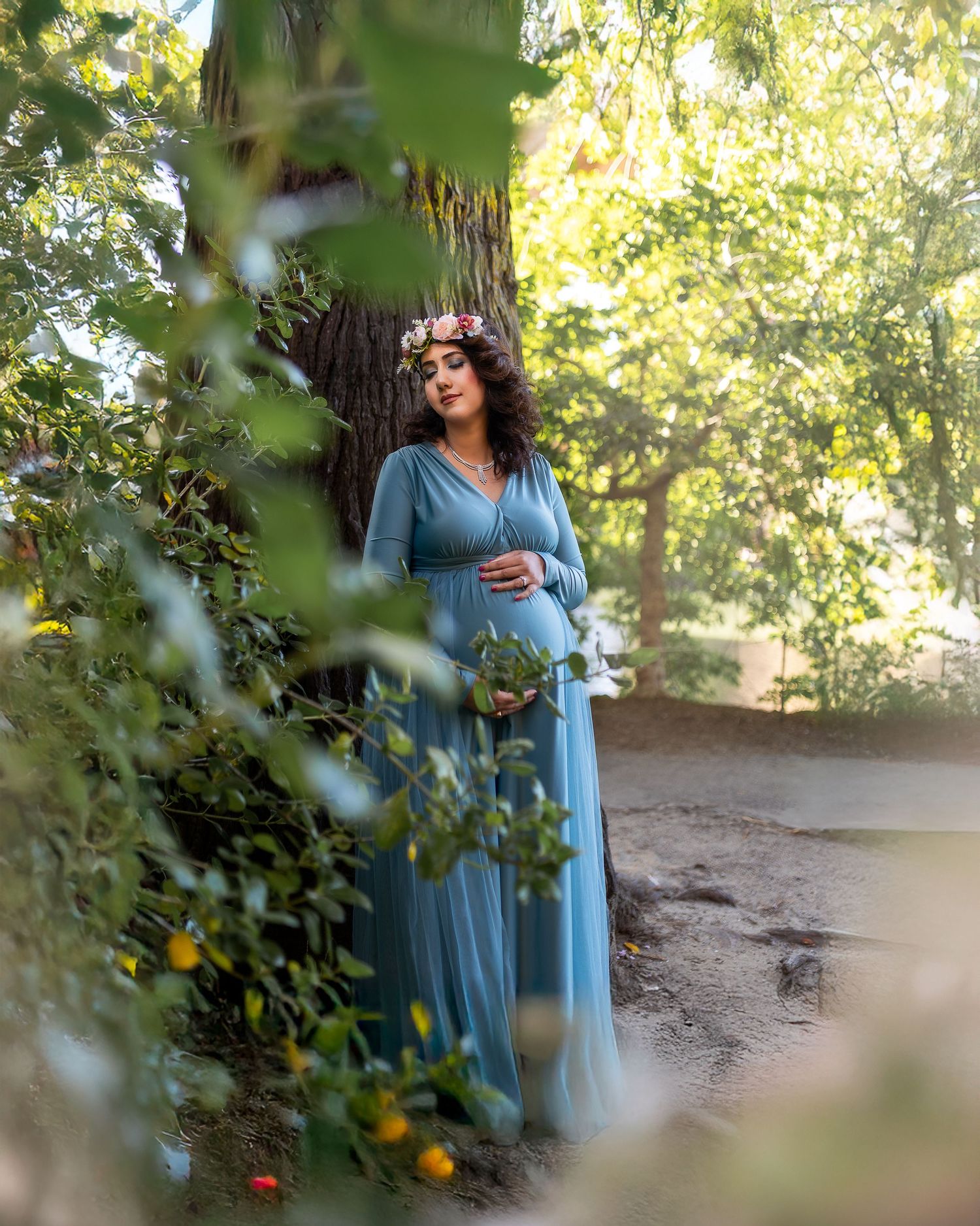 A person in a flowing light blue dress poses gracefully among sunlit trees and foliage in a natural outdoor setting.
