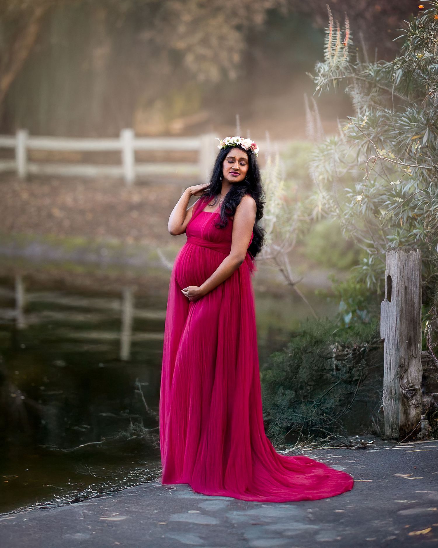 A person in a flowing magenta maternity gown poses outdoors near a white fence at sunset.