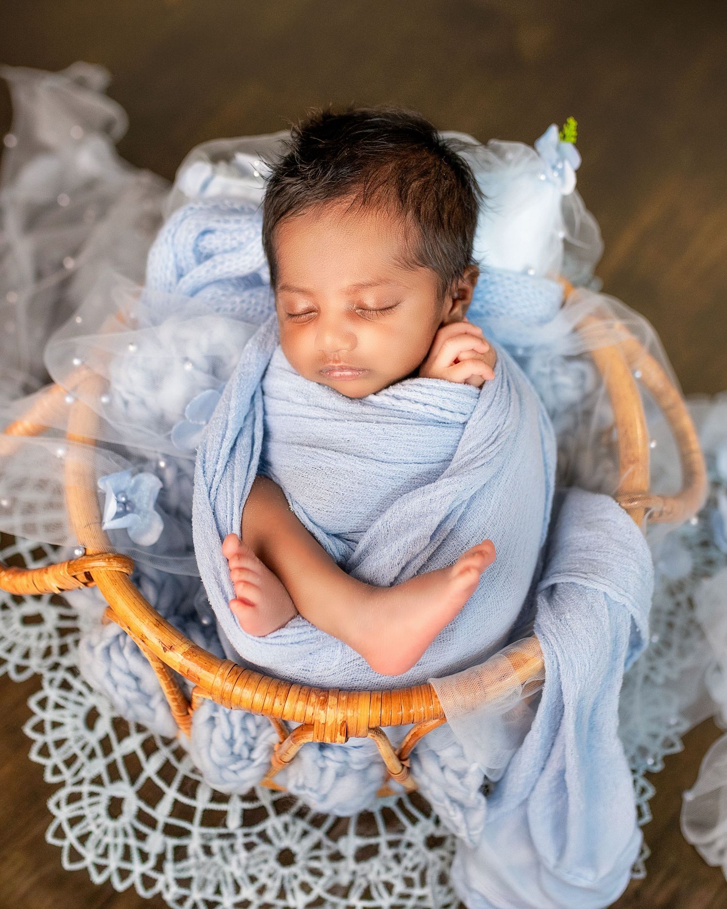 Newborn baby sleeping peacefully on white lace doily while wrapped in soft blue swaddling fabric.