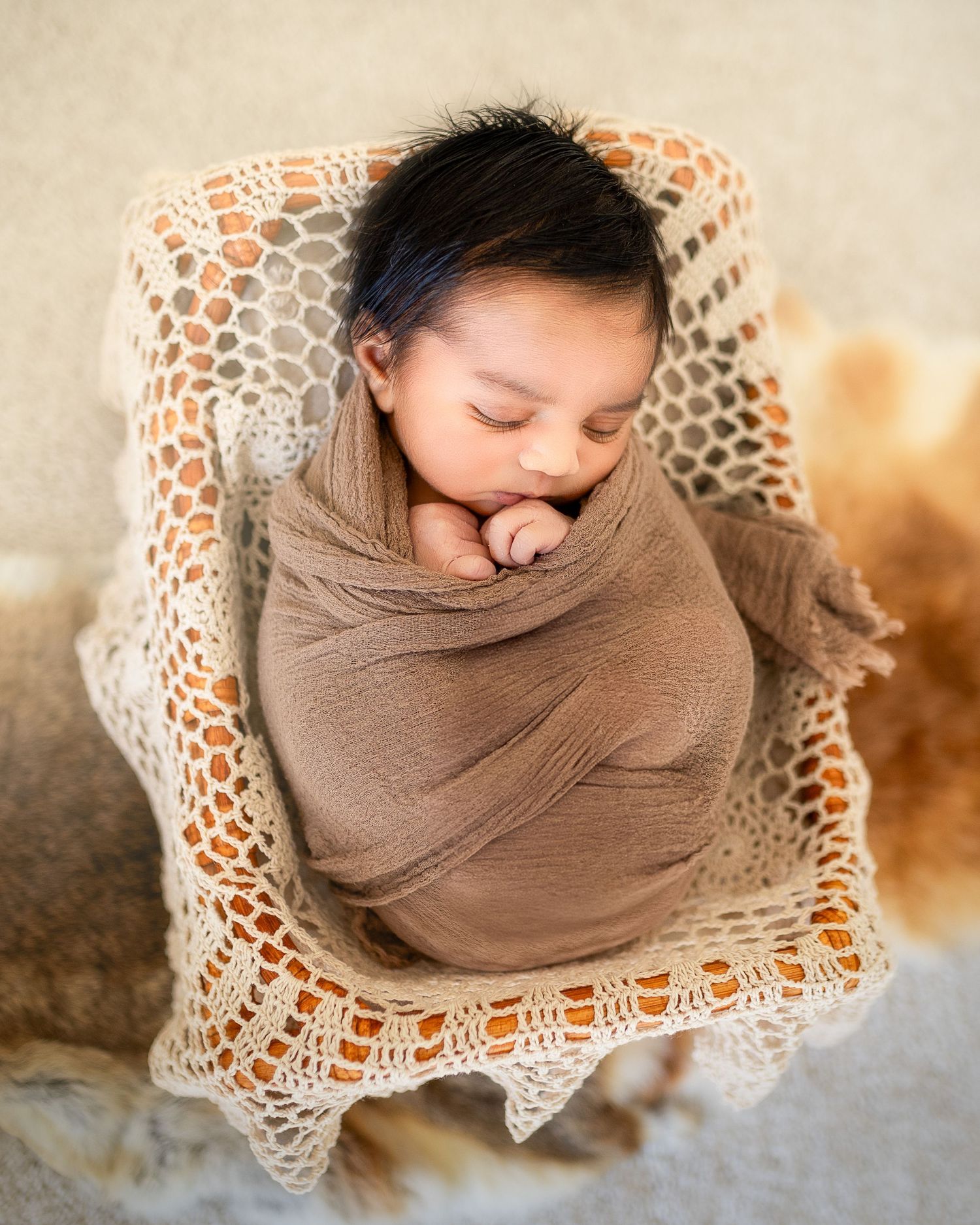 Overhead view of a peaceful newborn swaddled in brown fabric on a delicate crocheted blanket.