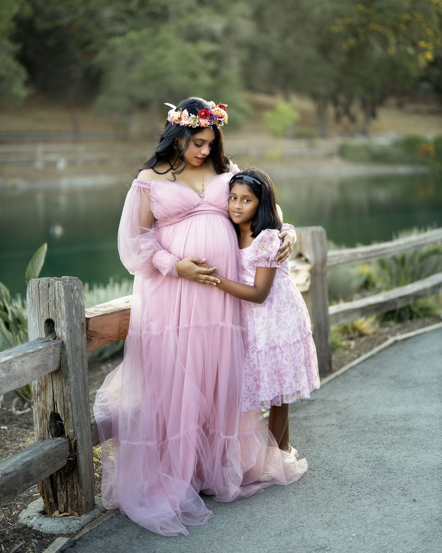 Two people in pink dresses share a tender moment on a wooden bridge by a serene lake.