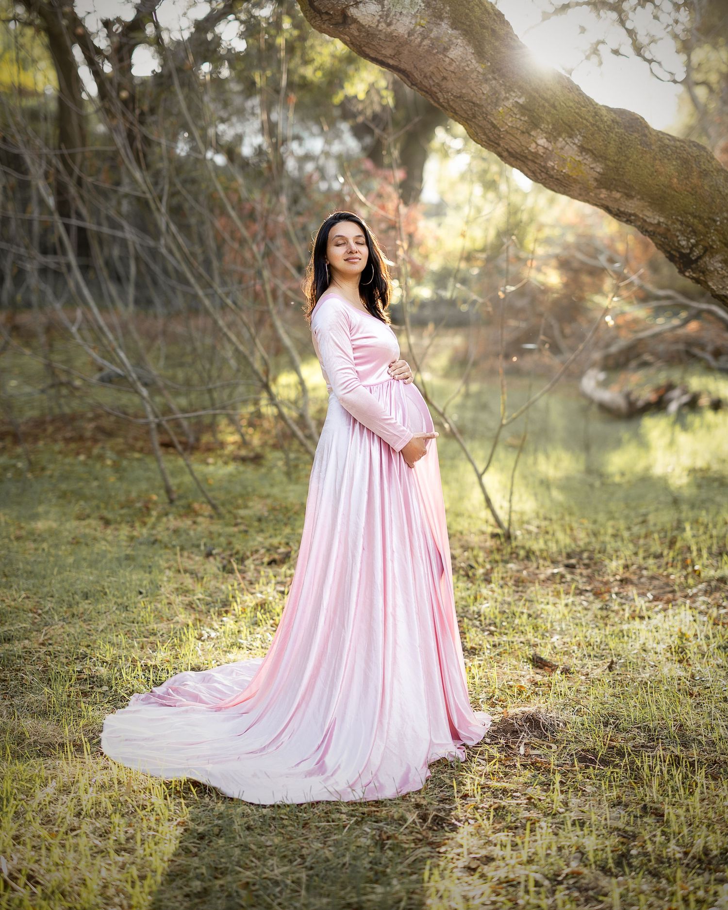 A person in a flowing pink maternity gown poses gracefully in a sunlit outdoor garden setting.