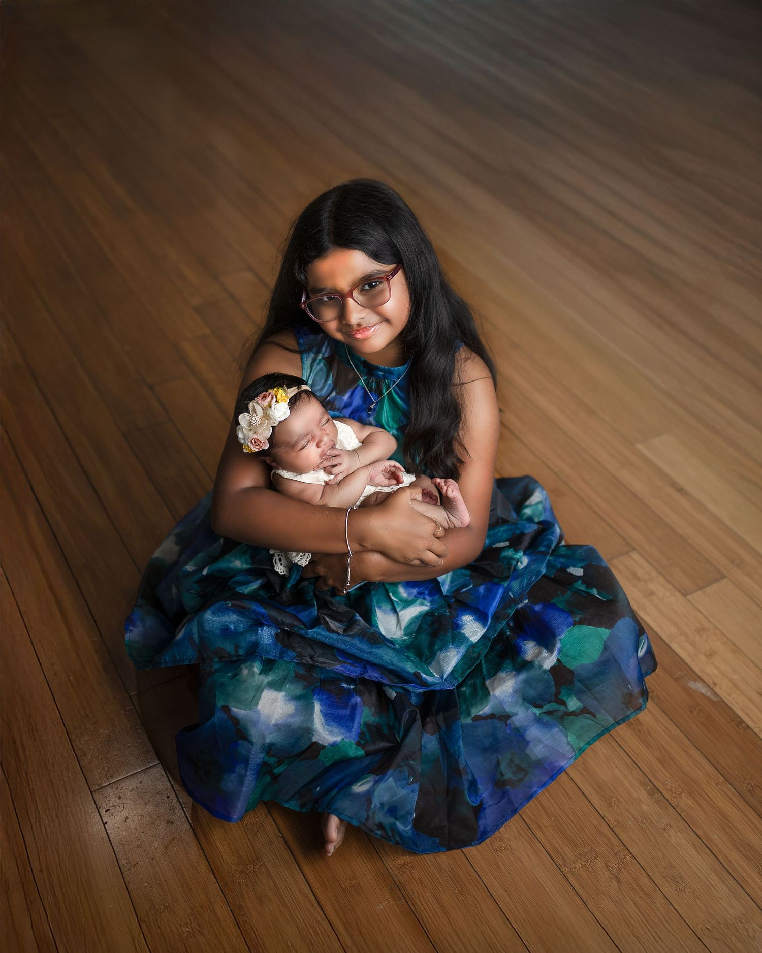 A young person in glasses wearing a blue floral dress holds a small baby while sitting on a hardwood floor.