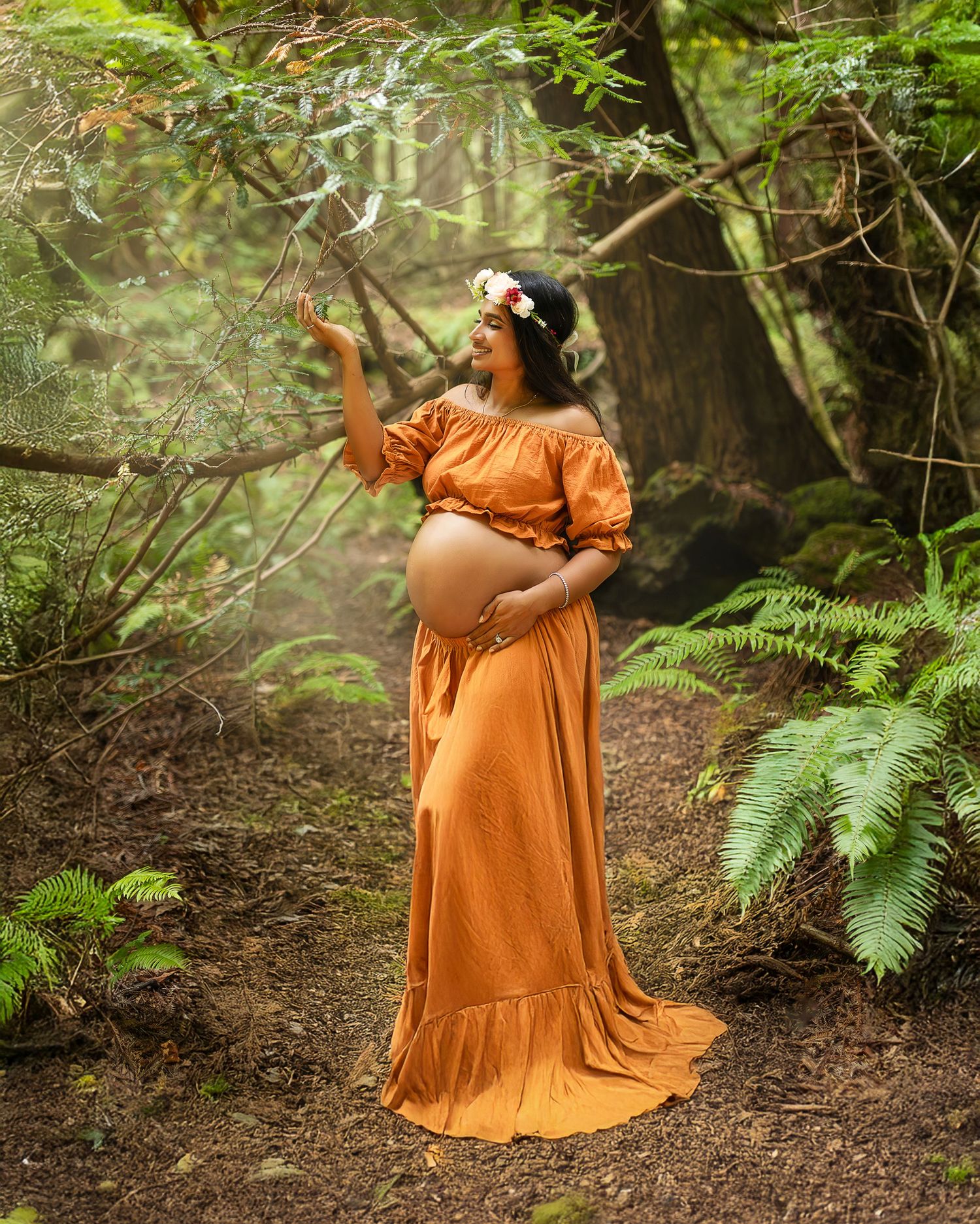 Expectant person in flowing orange dress poses in enchanted forest setting with ferns and filtered sunlight.