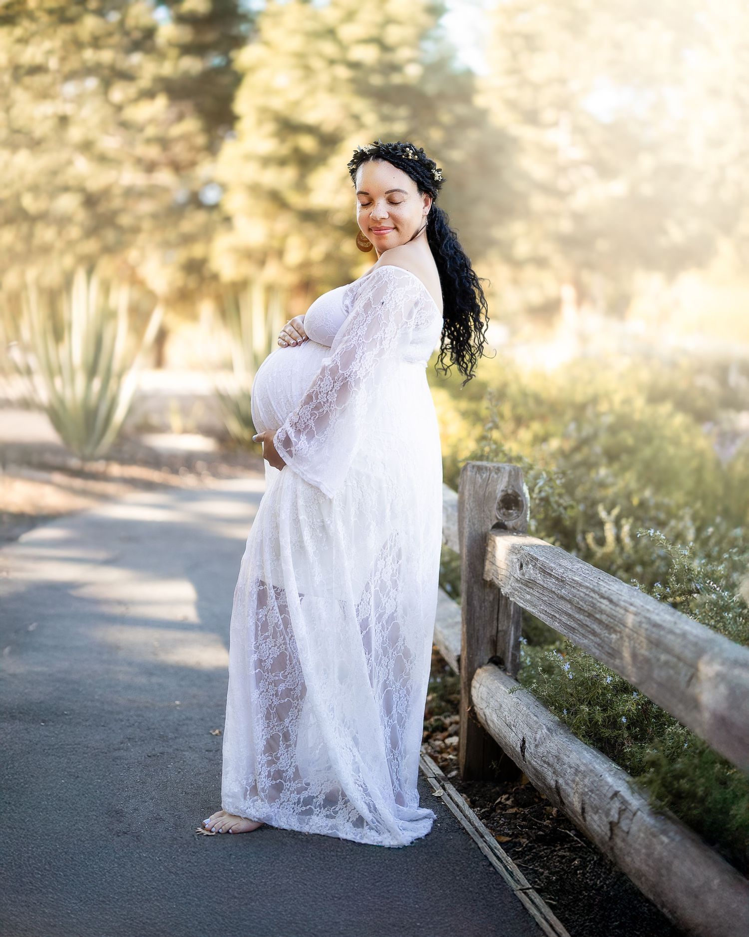 A figure in a flowing white lace dress poses gracefully against a rustic wooden fence in an outdoor setting.