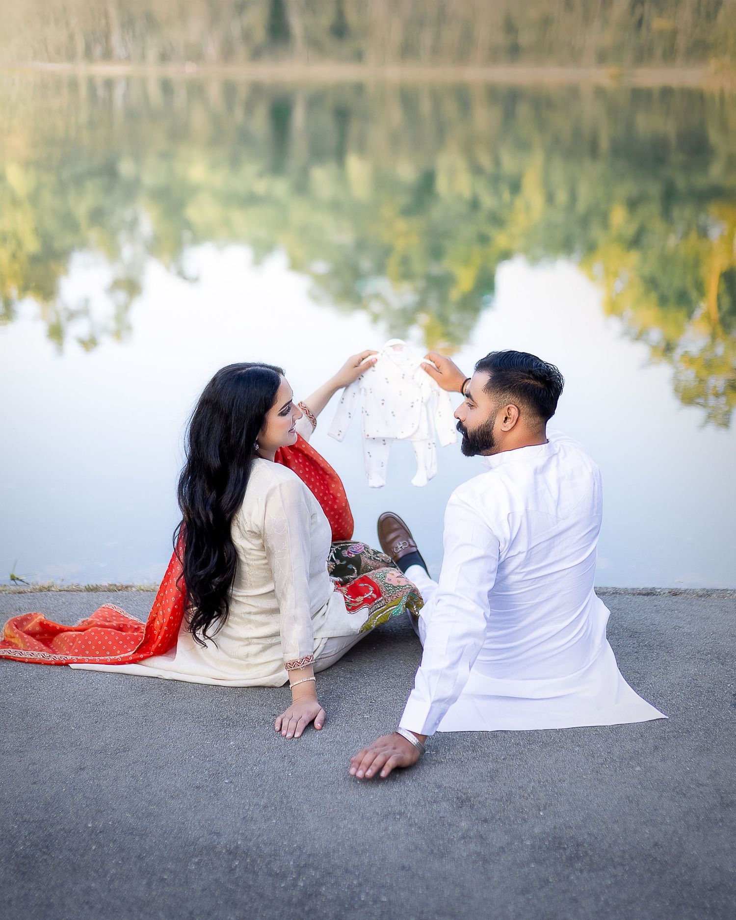 A couple lying on a stone surface near a lake enjoy a romantic moment in coordinated white and red outfits.