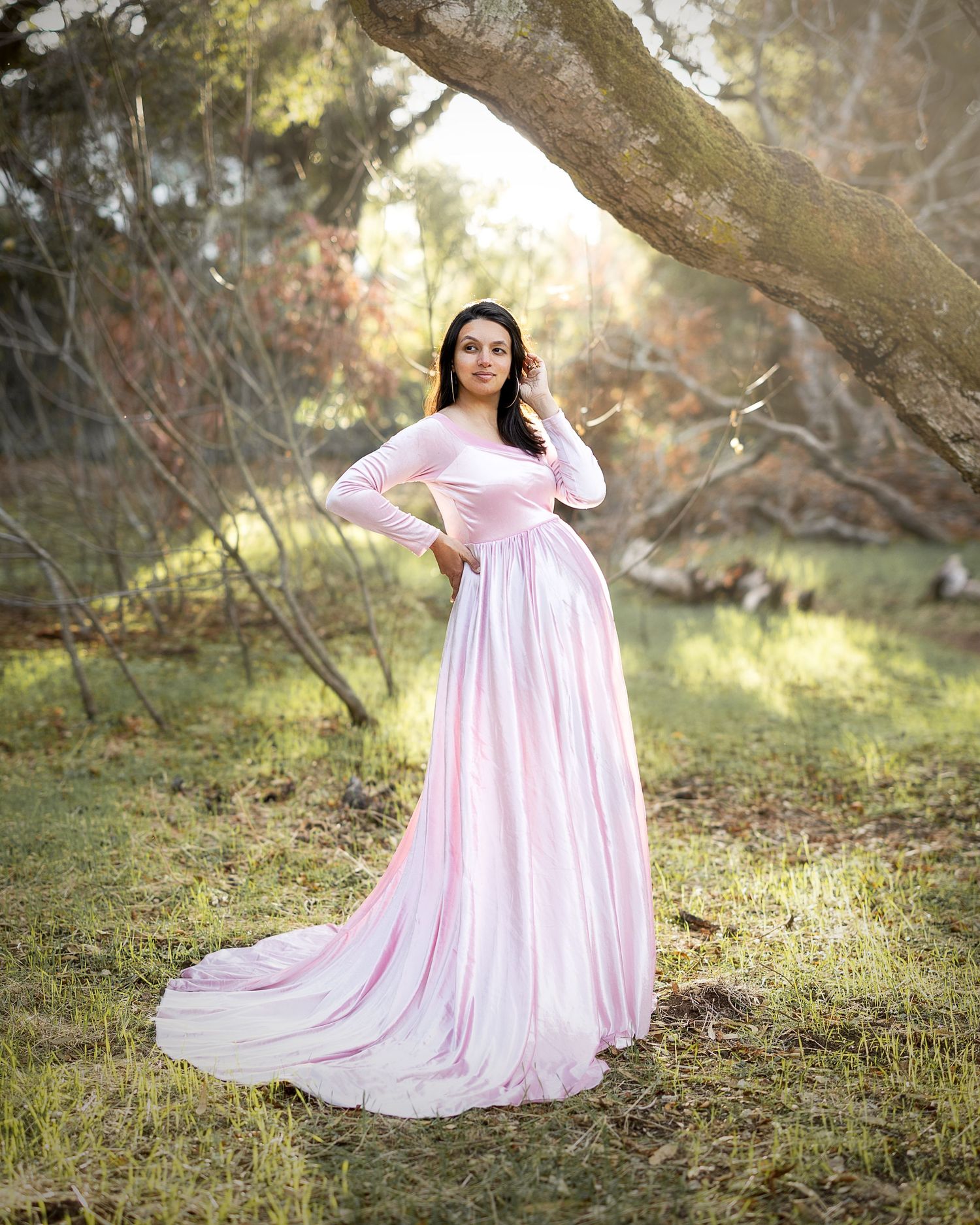 A person in a flowing pink gown poses gracefully in a sunlit woodland setting during a dreamy outdoor photo session.