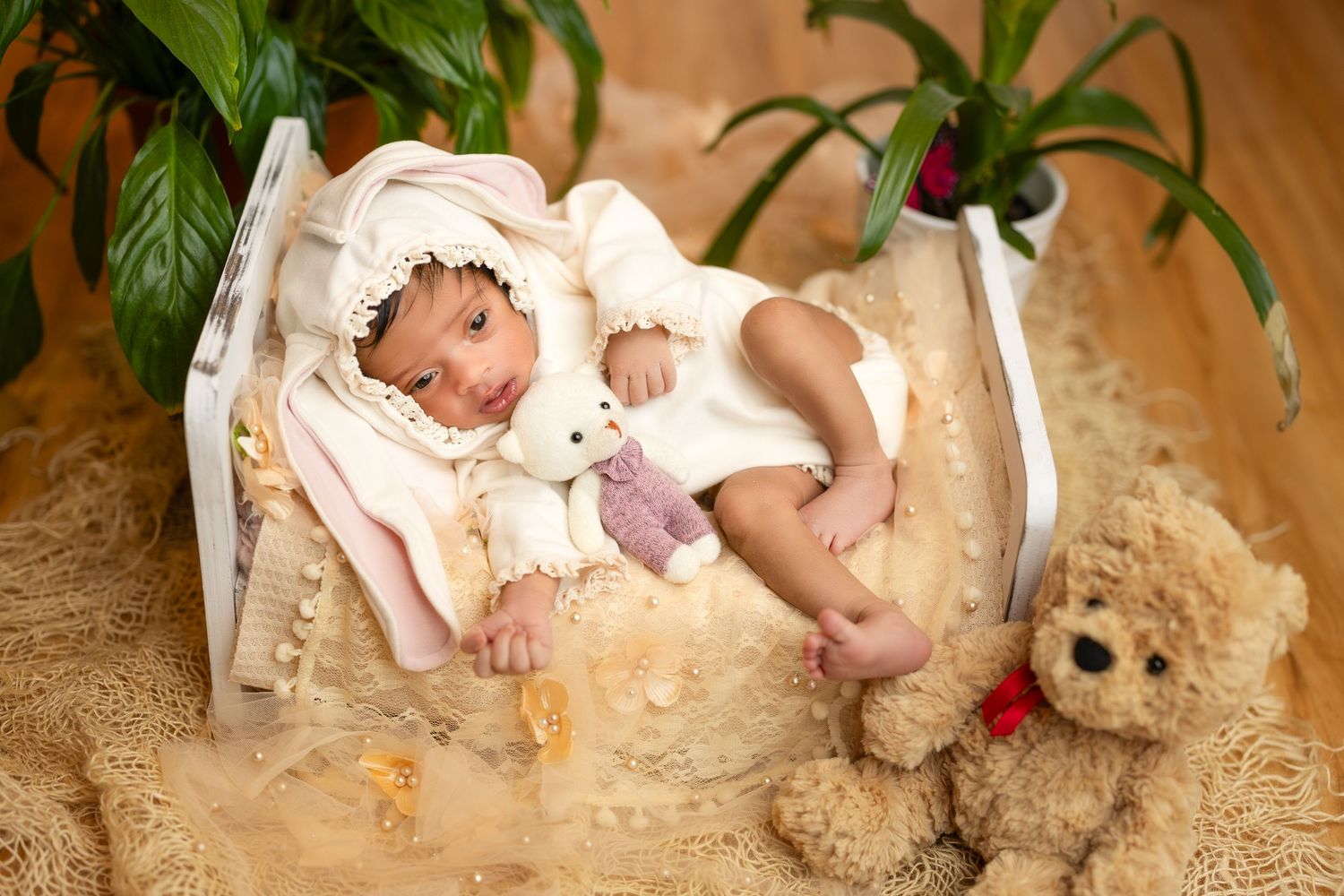 A baby lies on a cream-colored blanket surrounded by green plants in a natural light setting.