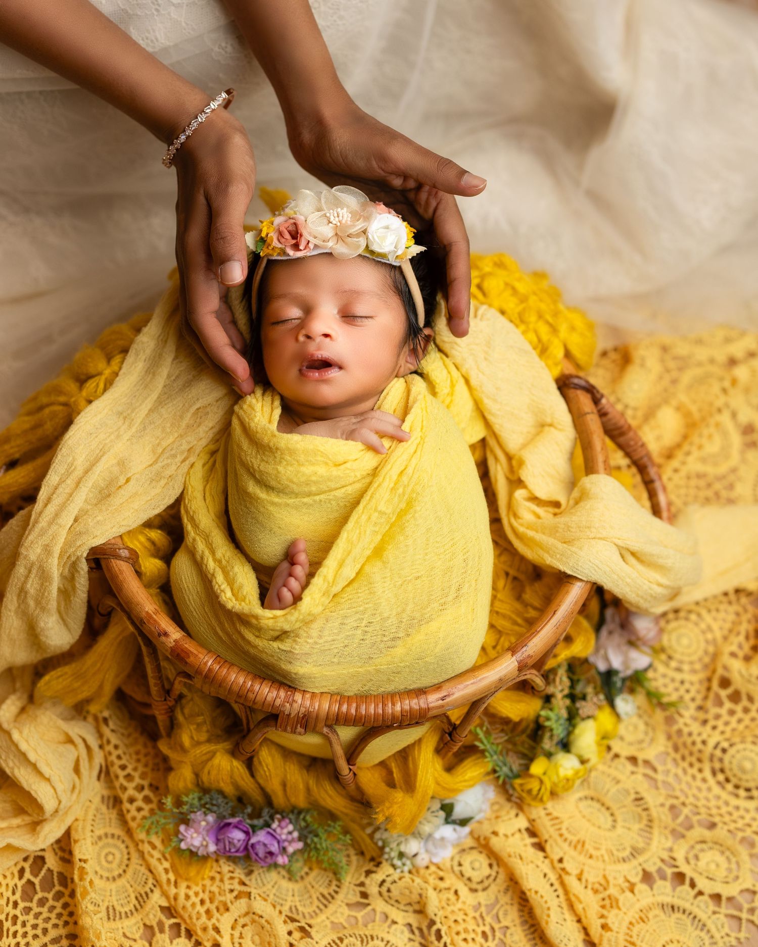 A newborn baby wrapped in soft yellow fabric rests peacefully in a woven basket with floral accents.