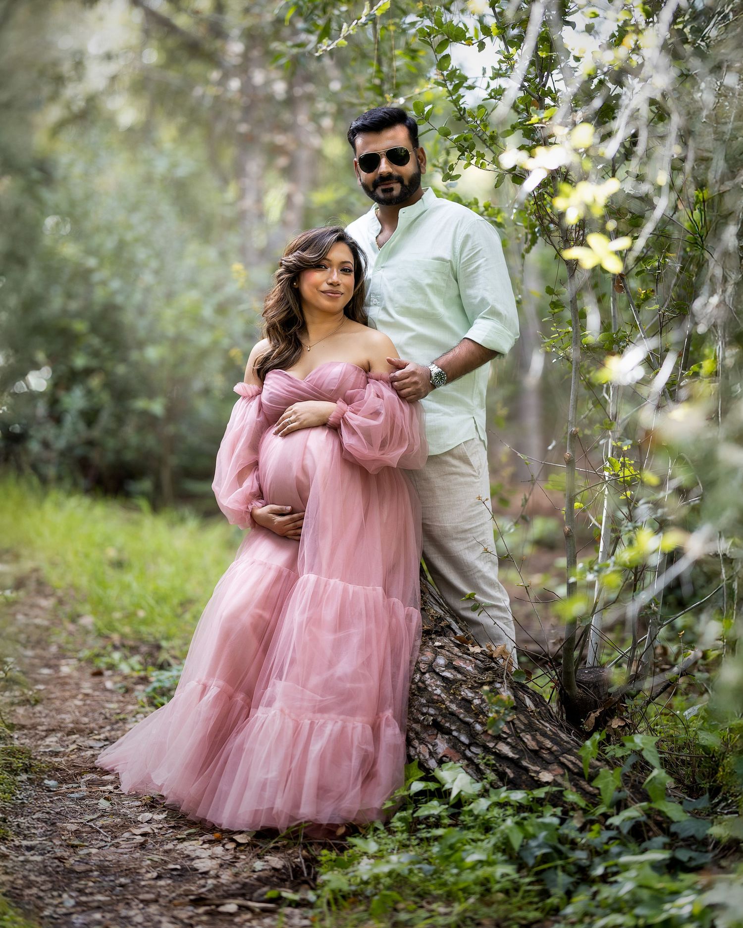 A couple poses in a dreamy outdoor setting with the person in a flowing pink maternity gown against lush greenery.