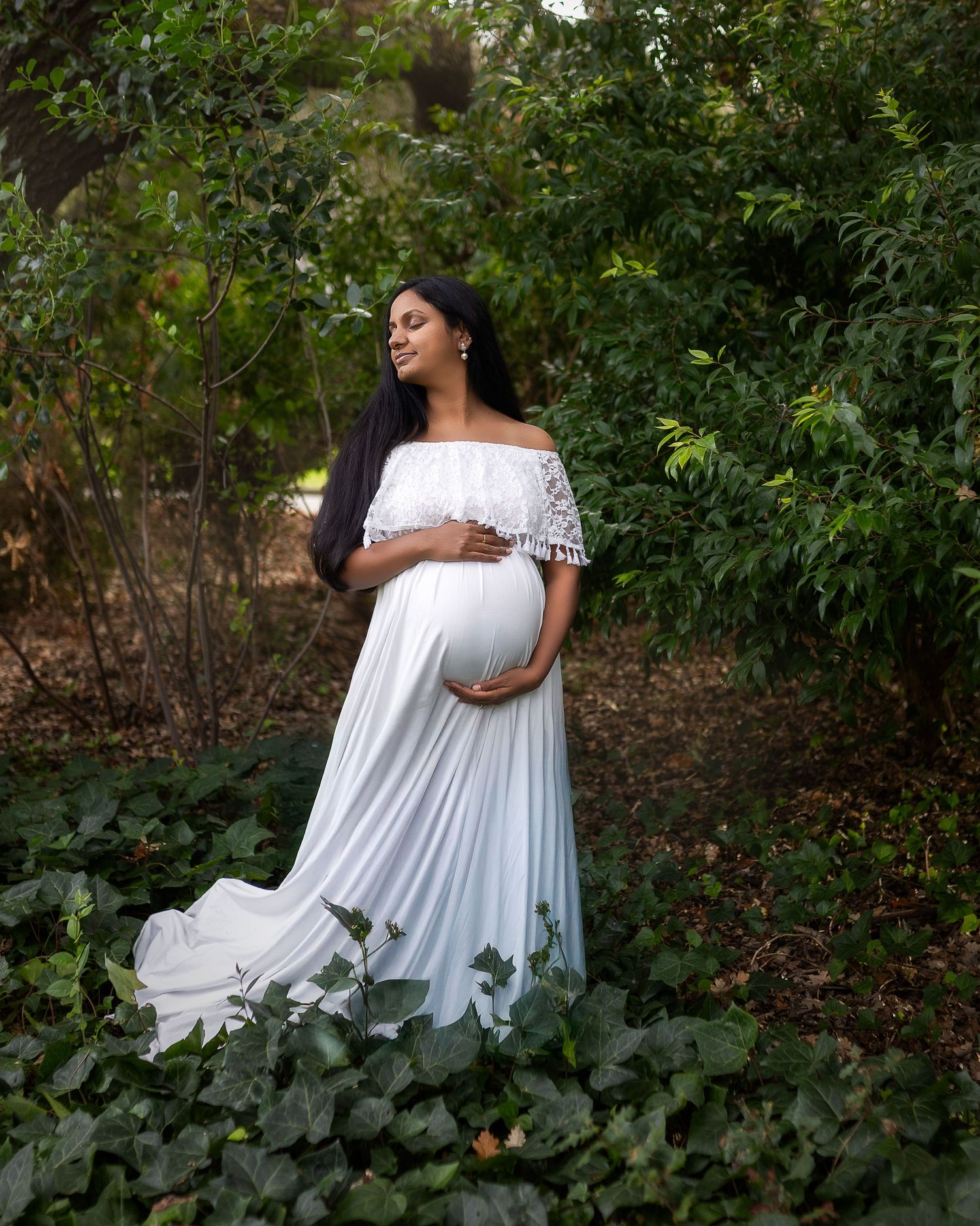 A maternity photoshoot in a lush garden setting captures someone in a flowing white off-shoulder dress among greenery.