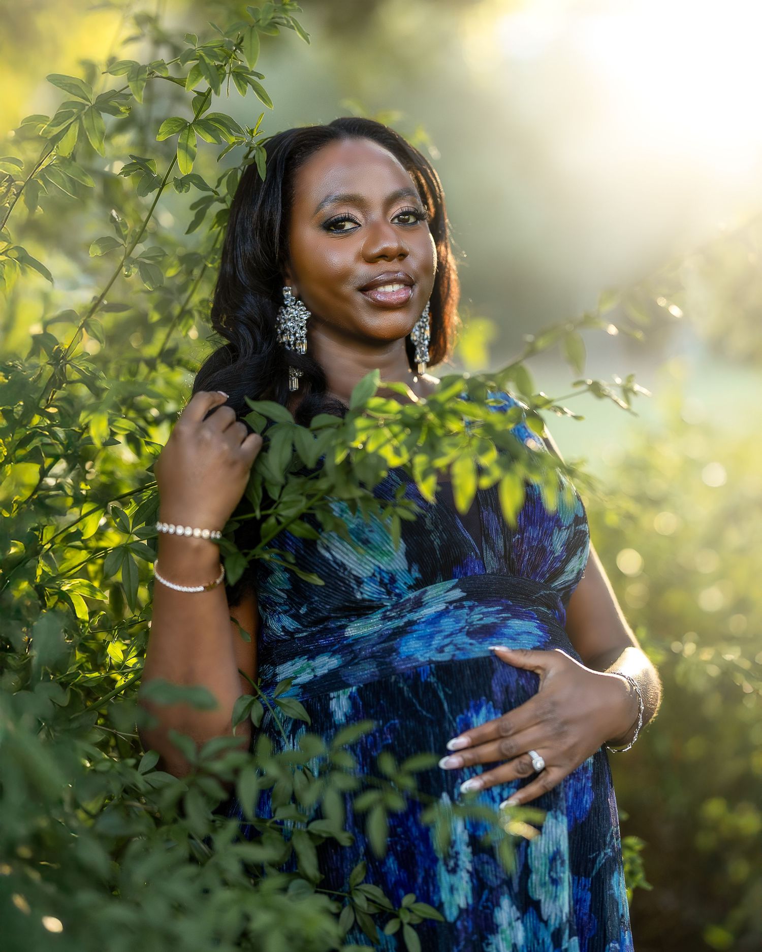 A woman in a blue tie-dye dress poses among green foliage during golden hour with sparkly earrings and bracelets.