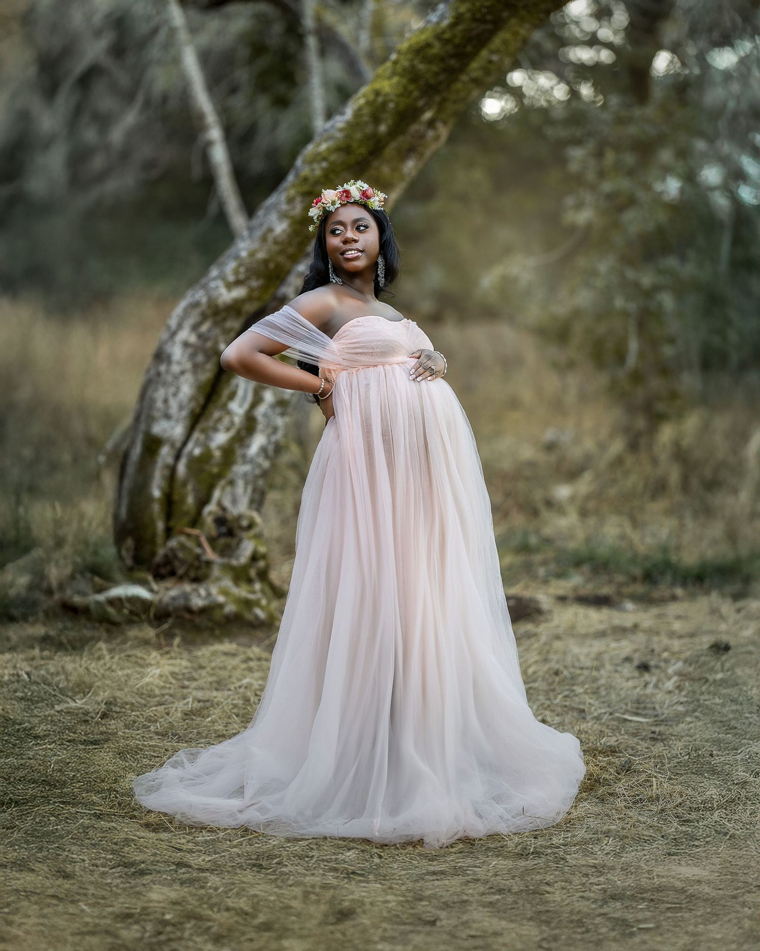 A flowing pink tulle maternity gown and floral crown create an ethereal woodland portrait in soft natural lighting.