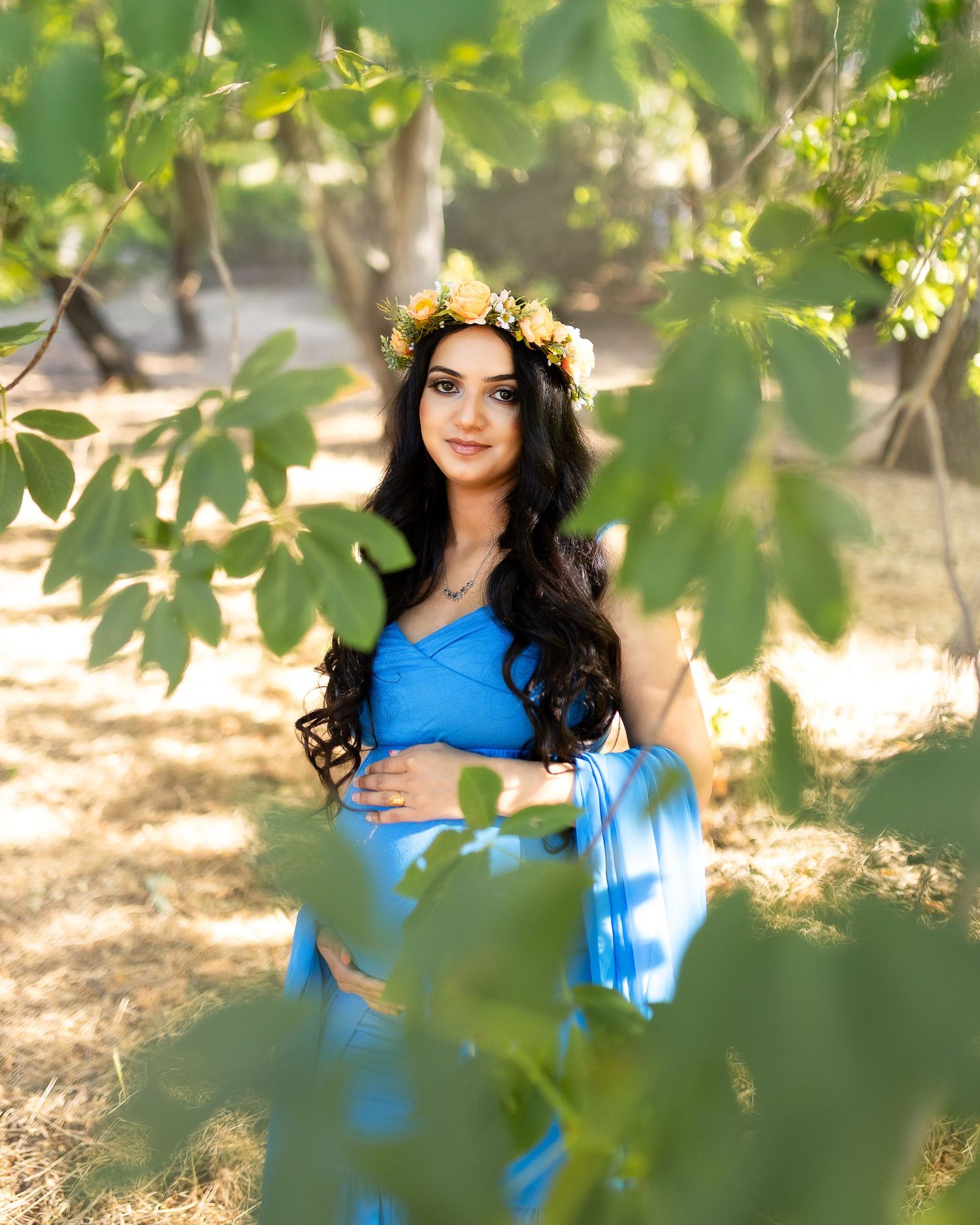 A person in a bright blue dress and floral crown poses gracefully among green foliage in an outdoor setting.