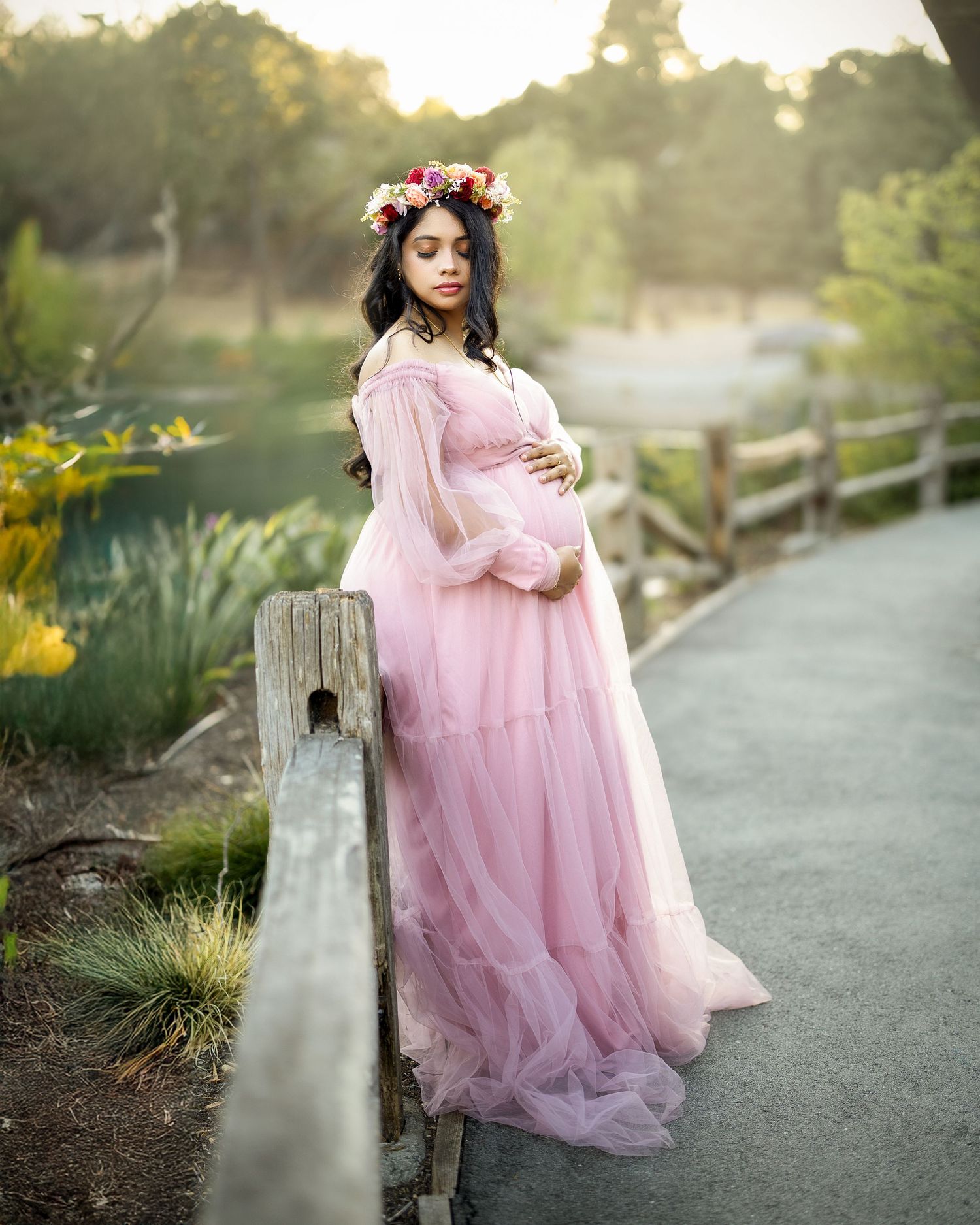 A dreamy maternity photoshoot in a pink flowing dress at sunset along a garden pathway.