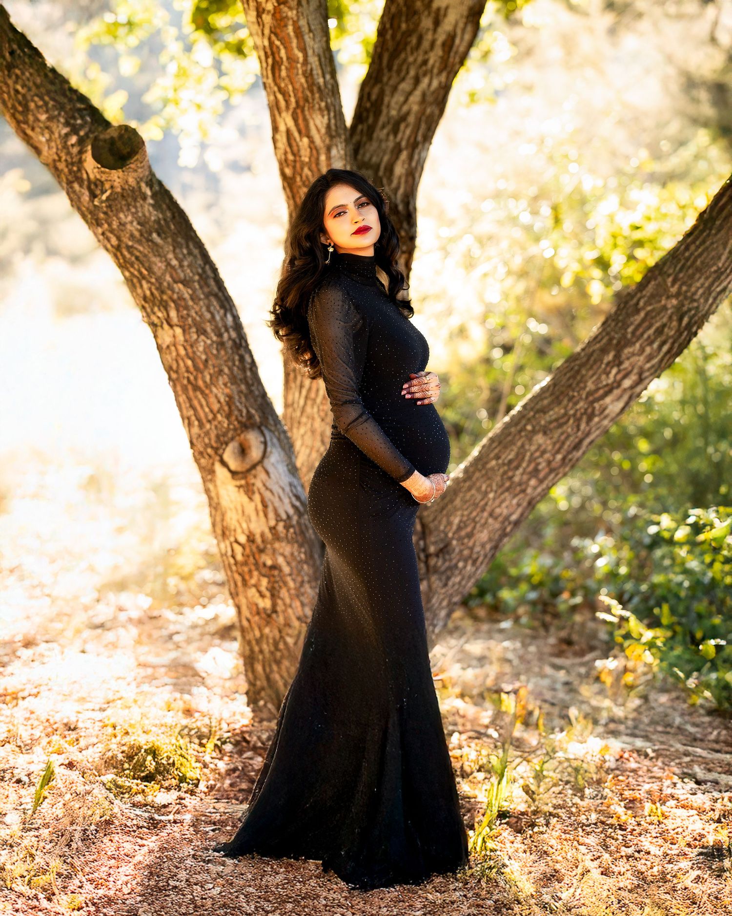 A figure in a flowing black dress poses gracefully against a tree trunk in a sunlit forest setting.