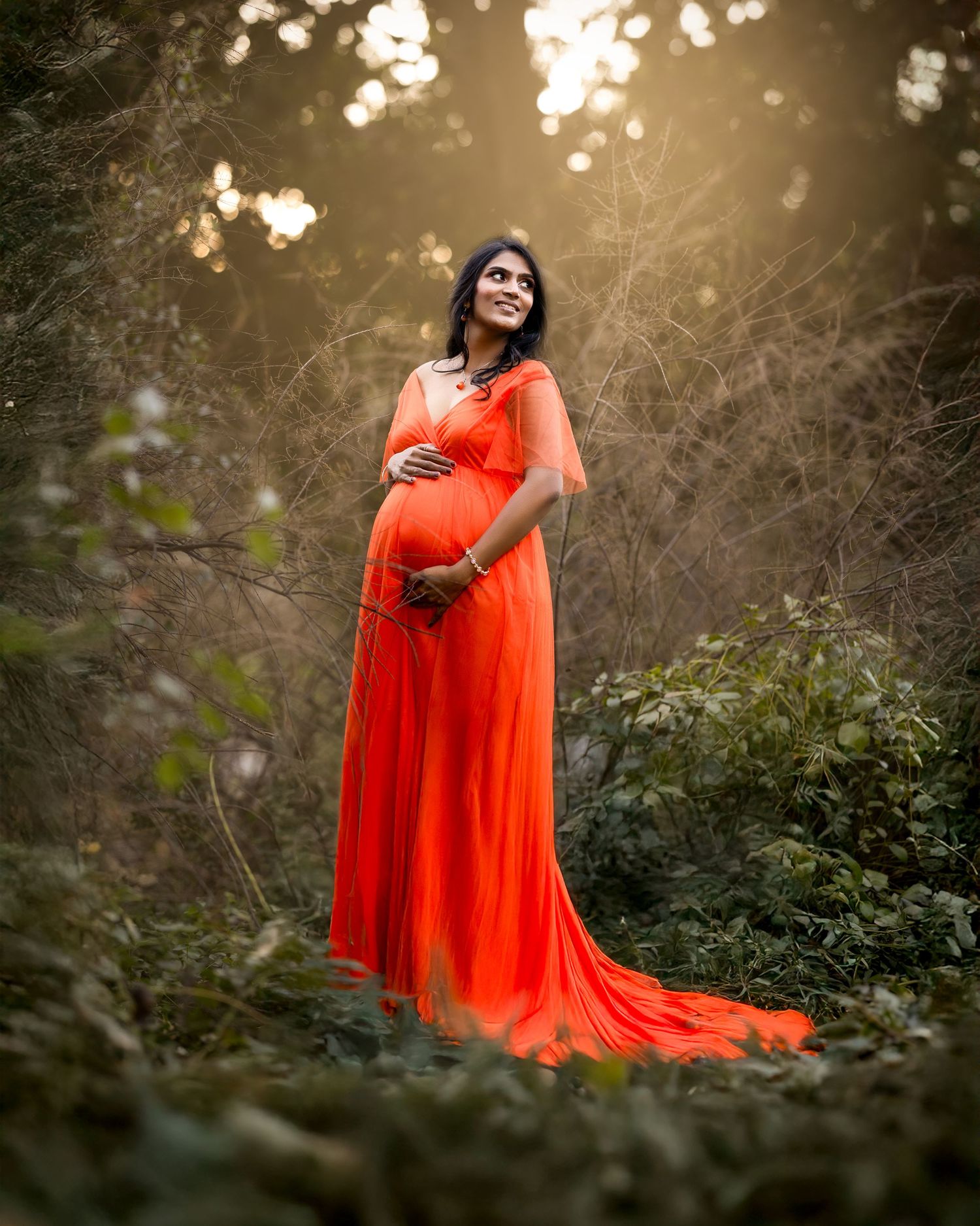 A maternity photo of an expectant mother in a flowing orange dress standing in a dreamy outdoor setting.