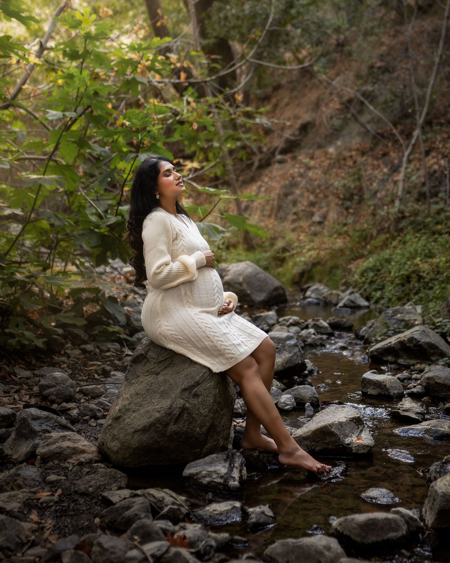 A person in a cream sweater relaxes on rocks by a peaceful stream in a wooded setting.