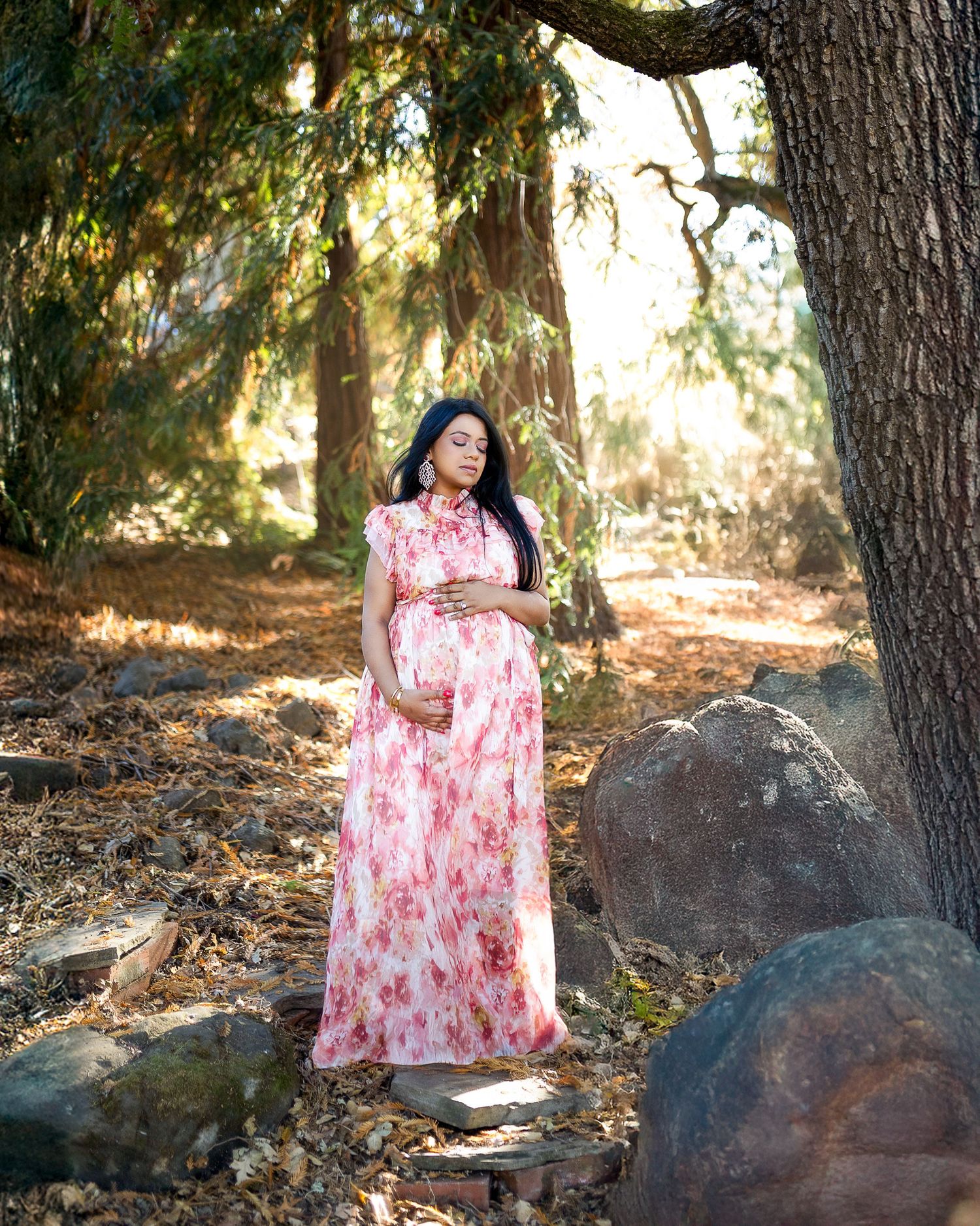 A person in a flowing pink floral maxi dress poses outdoors under a tree with large rocks in a natural setting.