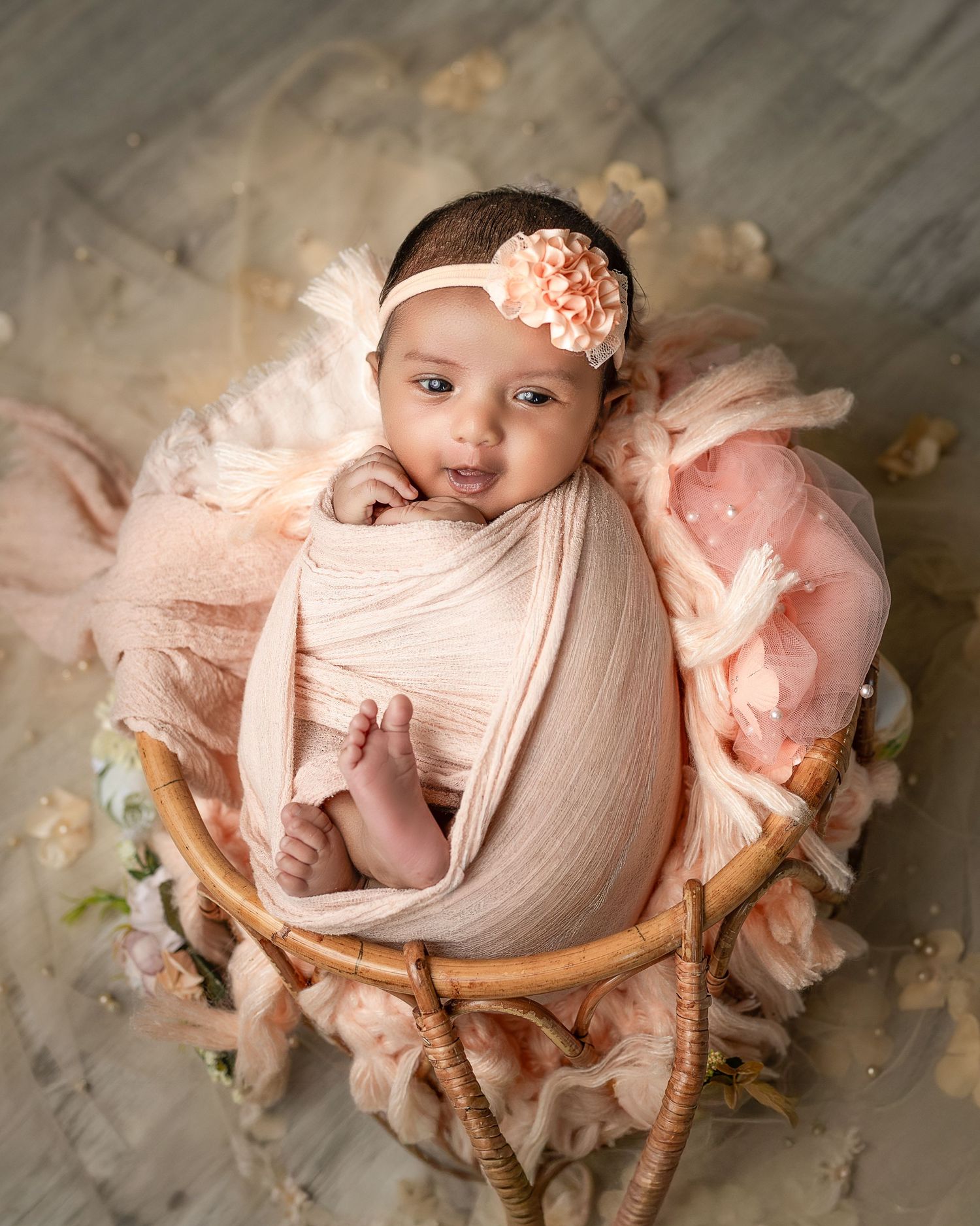 A newborn baby wrapped in soft pink fabric lies in a rustic wooden basket with delicate floral accents.