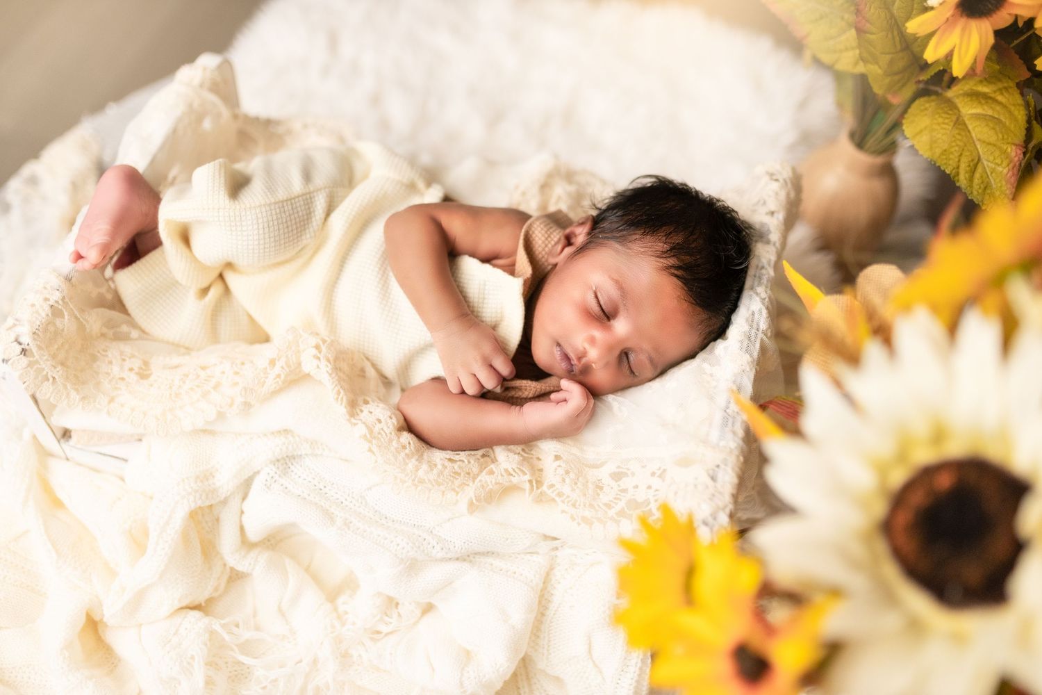 Newborn baby sleeping peacefully on white blanket surrounded by yellow sunflowers and autumn leaves.