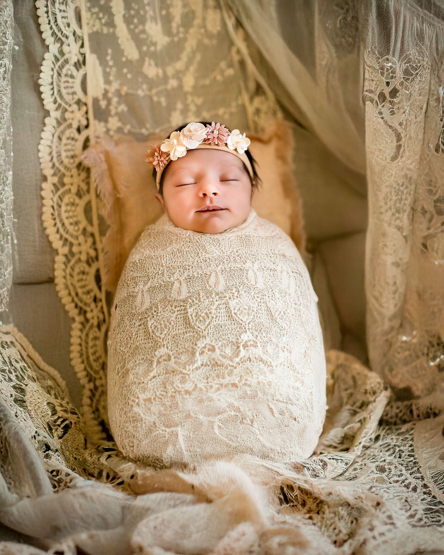 Sleeping newborn wrapped in delicate white lace with a floral crown against vintage lace curtain backdrop.