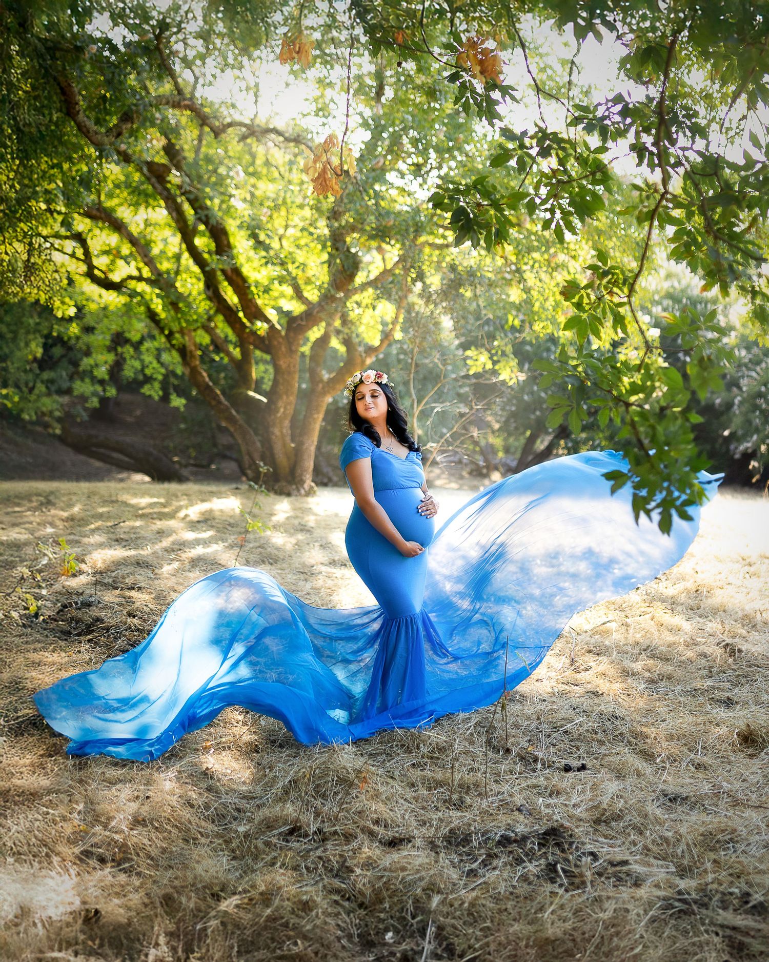 A flowing blue dress billows gracefully in the wind against a natural outdoor setting with dappled sunlight through trees.