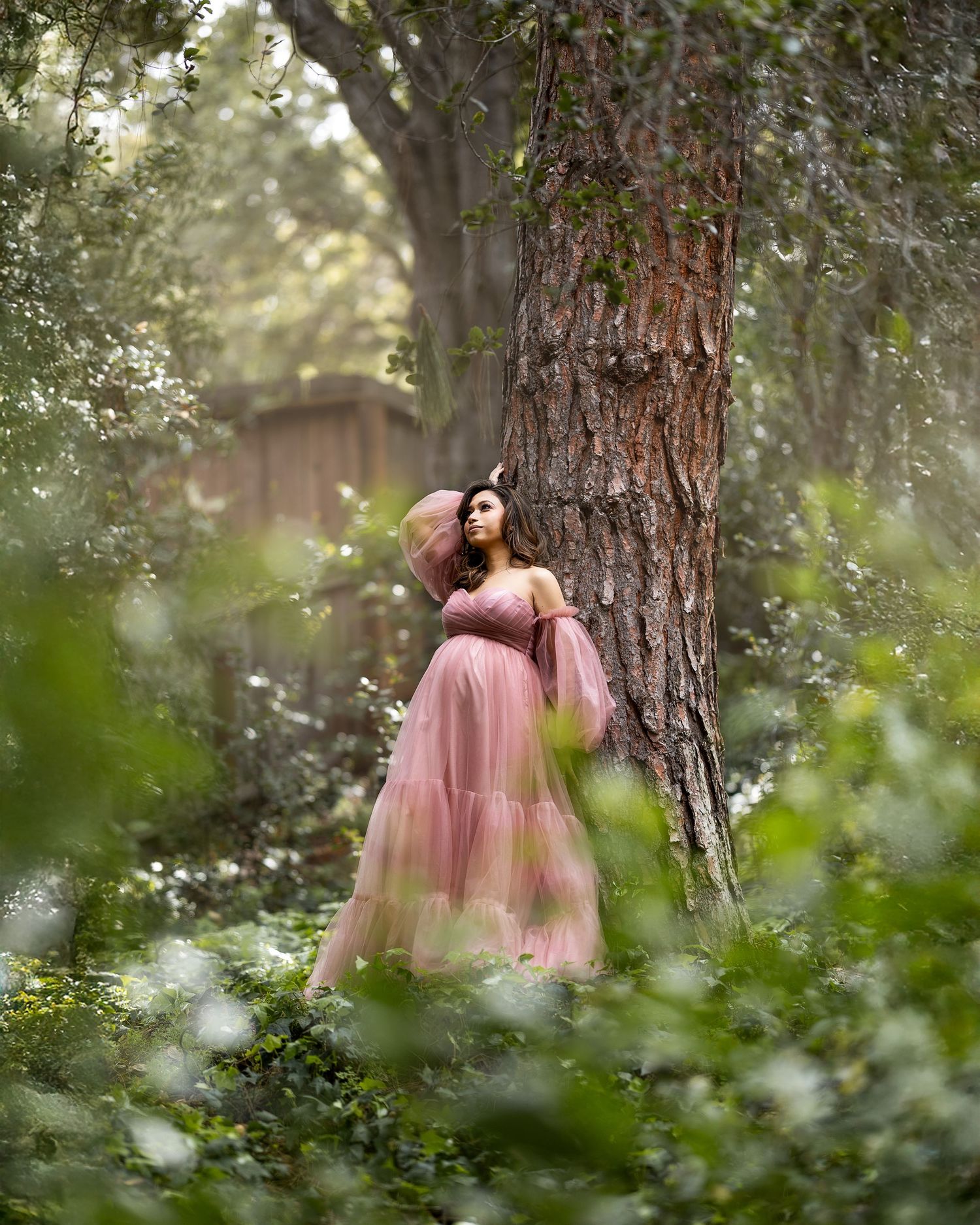 A romantic couple in pink dresses shares an intimate moment among tall pine trees in a dreamy forest setting.