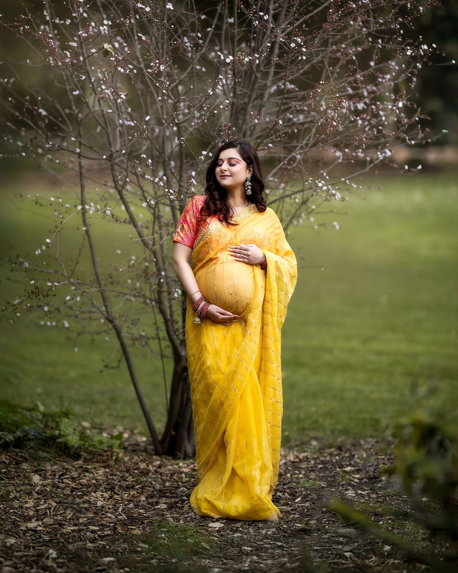 A figure in a vibrant yellow sari poses gracefully among blooming cherry blossom branches in a garden setting.