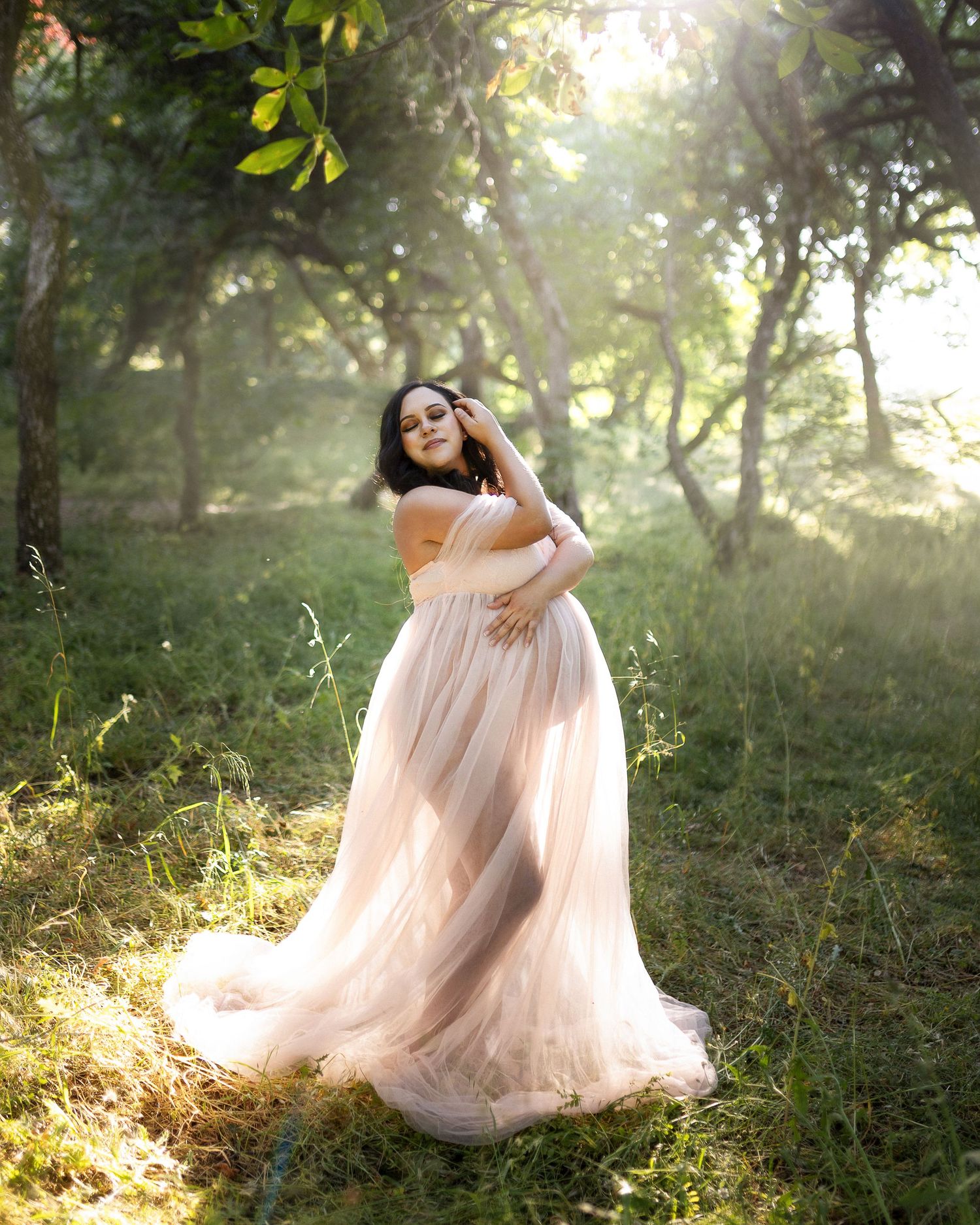 A flowing blush tulle gown catches golden sunlight in a dreamy forest setting during a maternity photoshoot.