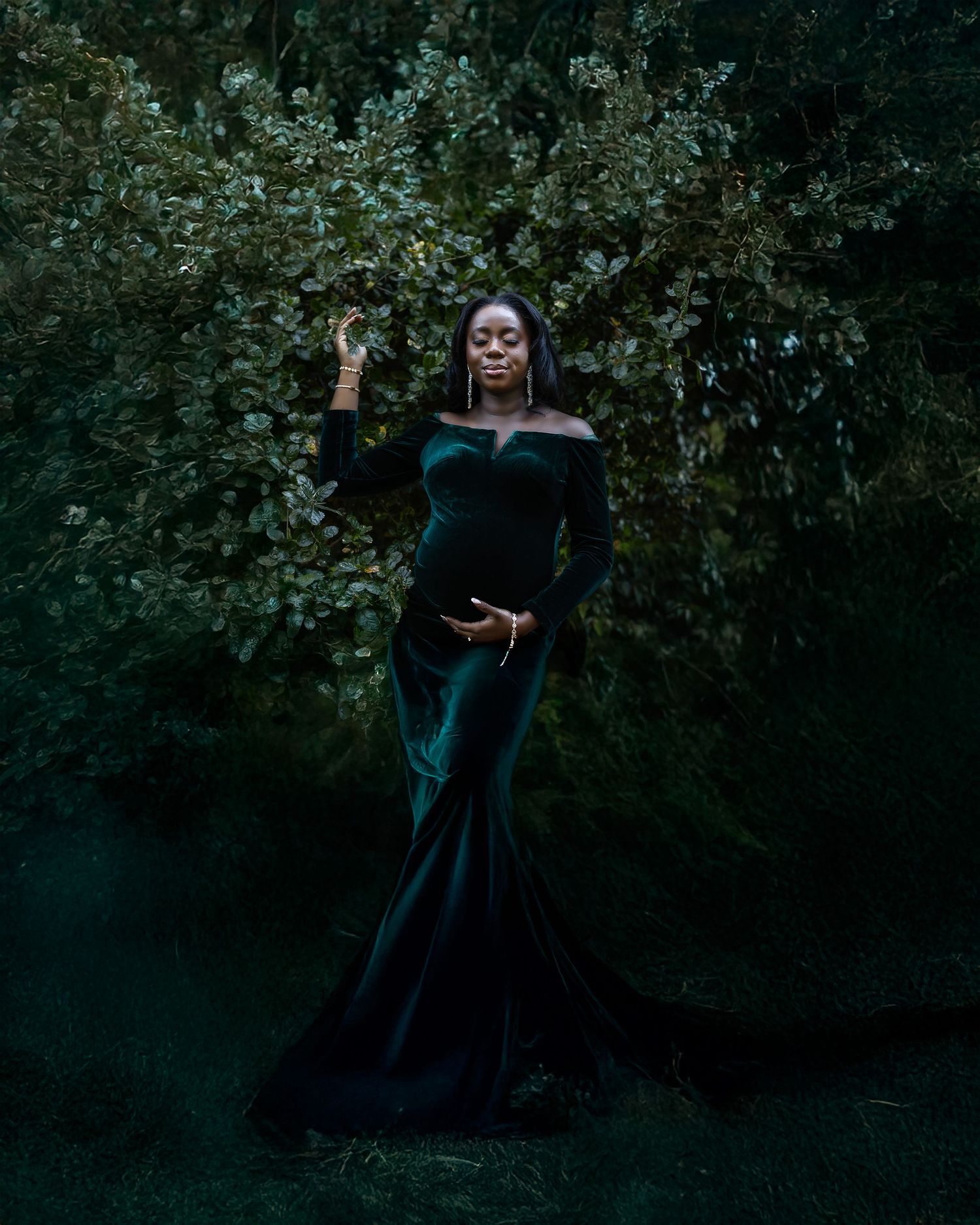 A flowing black evening gown is photographed against a dark, moody forest backdrop with dramatic lighting.