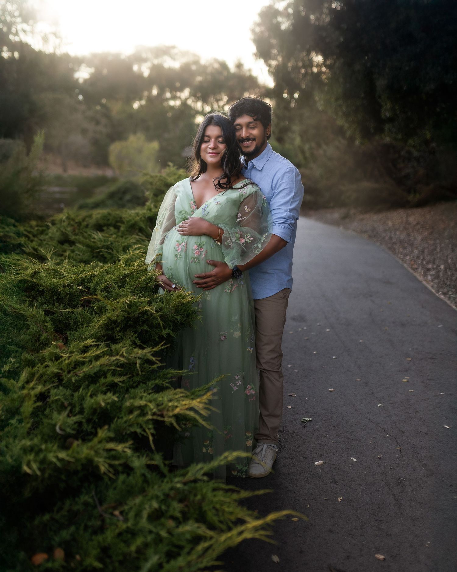 A couple embraces on a path during a maternity photoshoot at sunset with green foliage in the background.