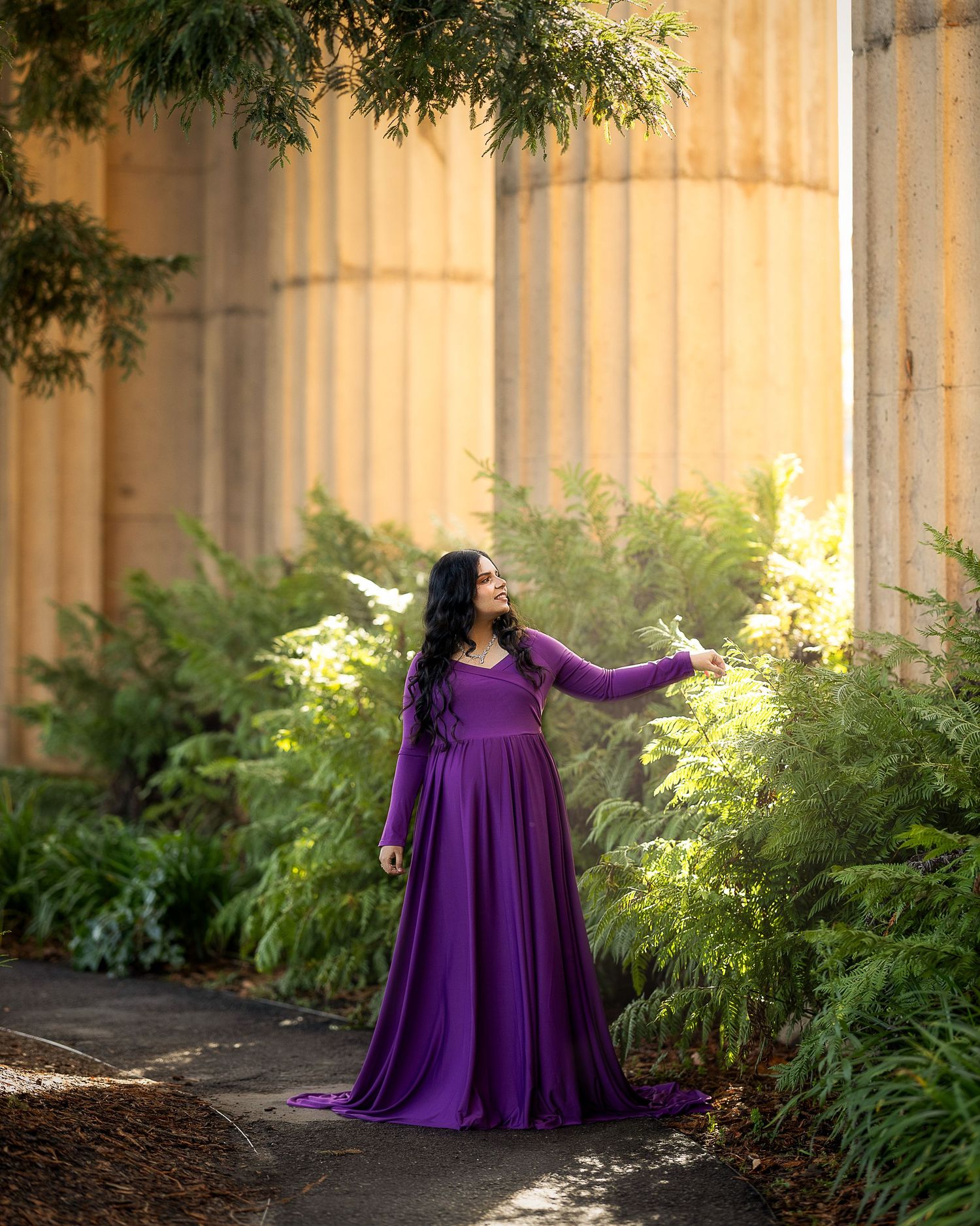 A person in a flowing purple dress poses gracefully in front of a sunlit wall surrounded by lush green ferns.