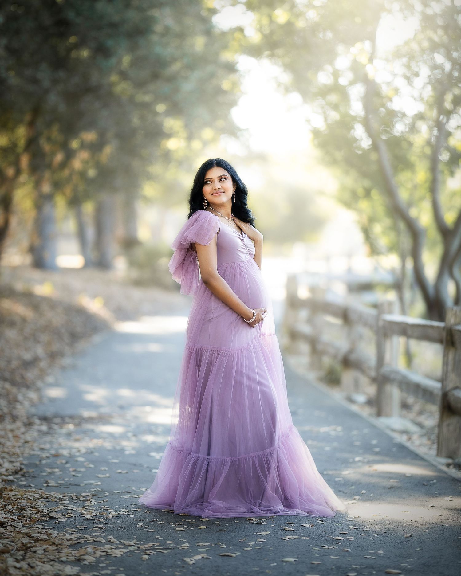 A person in a flowing lavender tulle gown poses gracefully on a sunlit tree-lined path during a dreamy outdoor photoshoot.