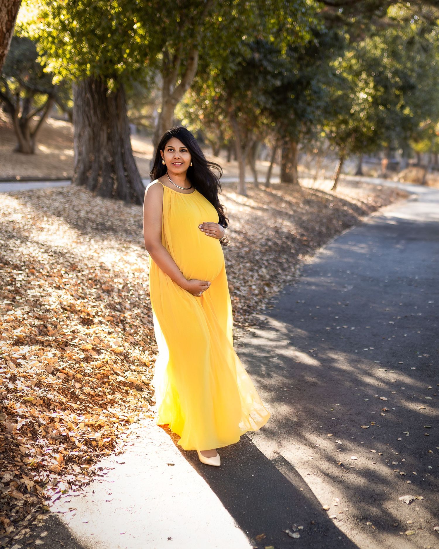 A person in a flowing yellow maternity dress poses on a tree-lined path during autumn with golden sunlight.