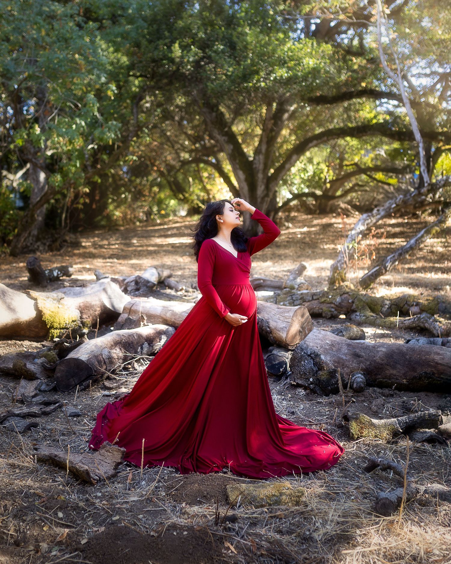 A flowing red maternity gown creates an enchanting scene against a wooded backdrop with sunlit trees and scattered rocks.