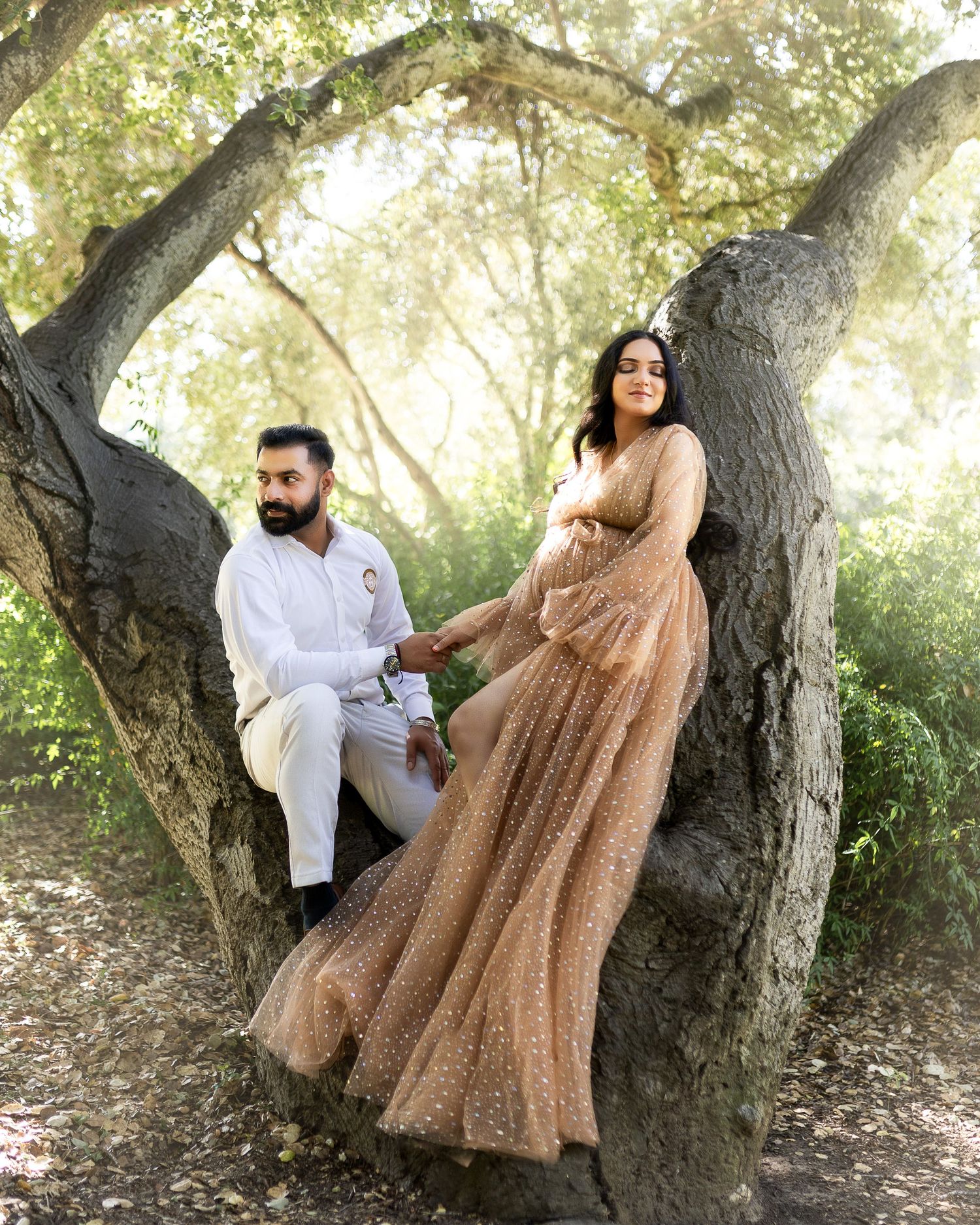 A couple in casual attire poses together near a large tree trunk in a sunlit outdoor setting.