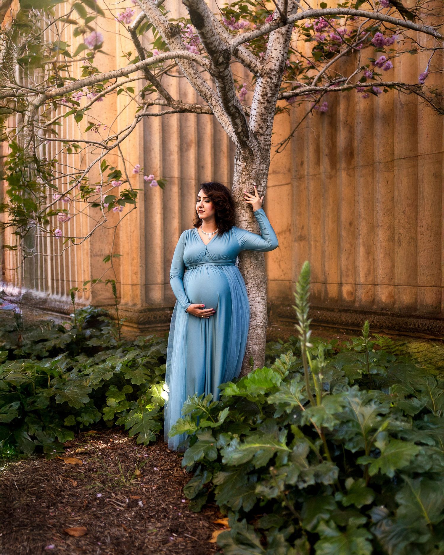 A light blue maternity dress flows elegantly in a garden setting with stone columns and autumn foliage.