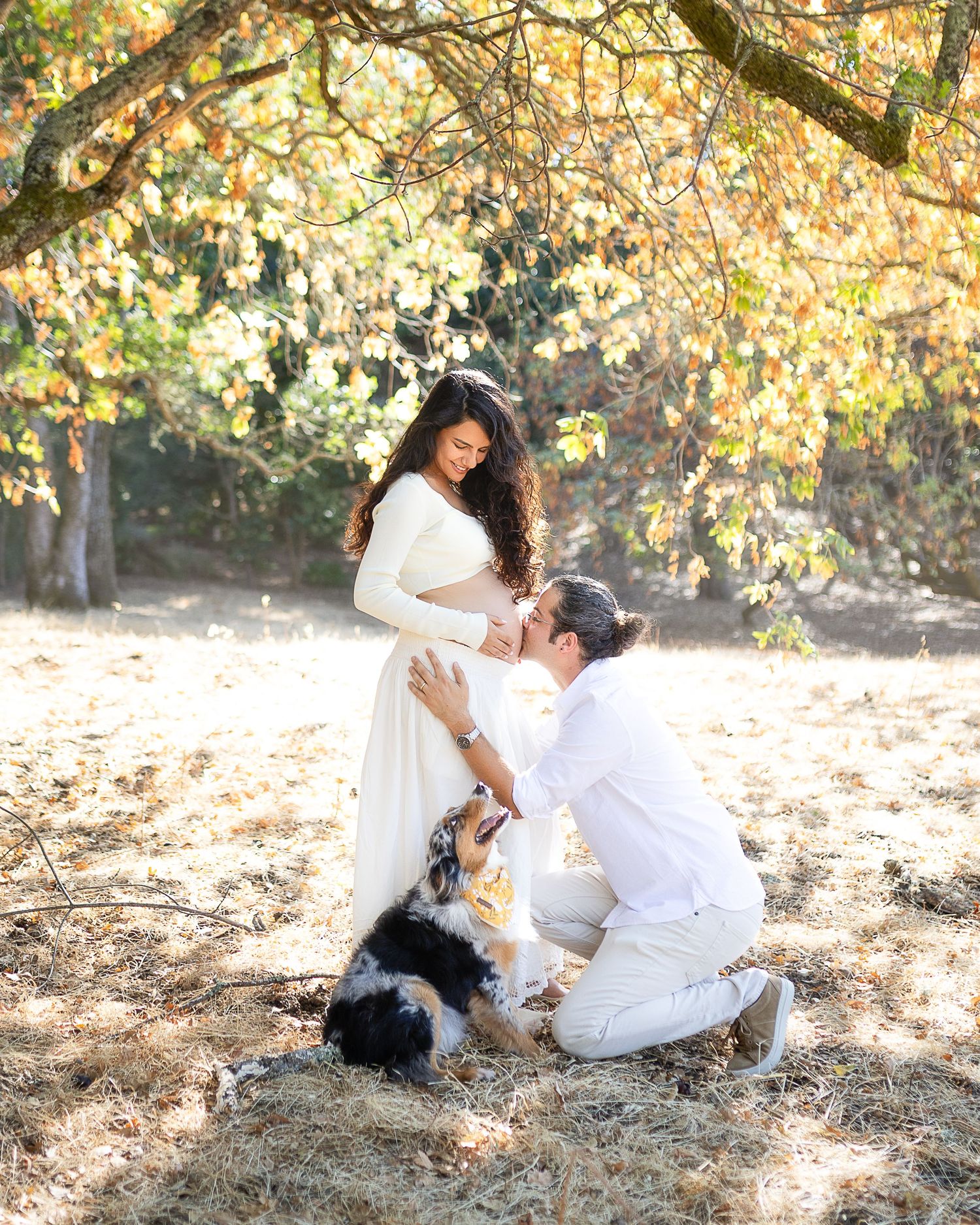 A couple in white outfits sits together on autumn ground surrounded by golden leaves and sunlit trees.