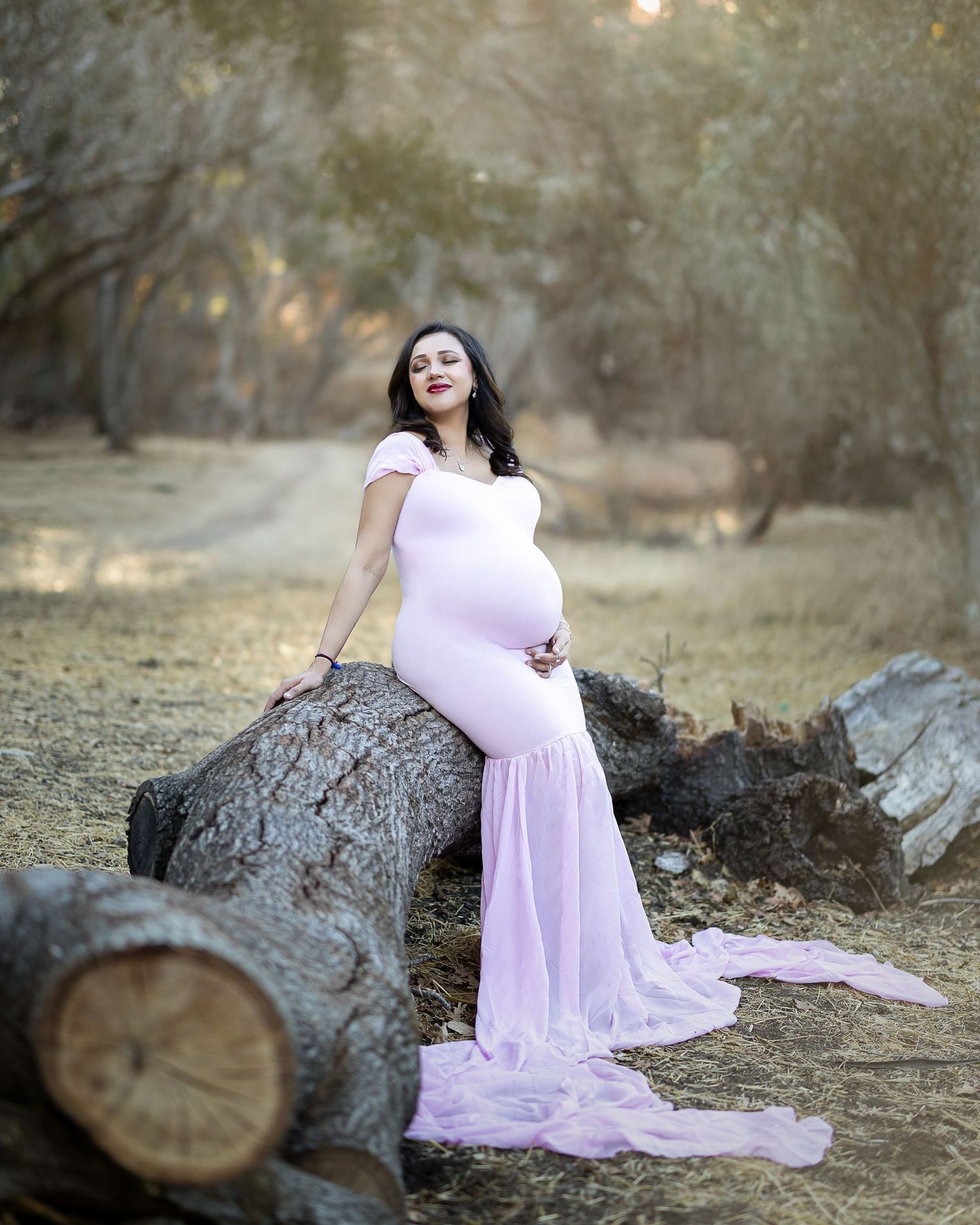 A flowing pink maternity gown creates an ethereal scene against a rustic woodland backdrop with fallen logs and muted lighting.